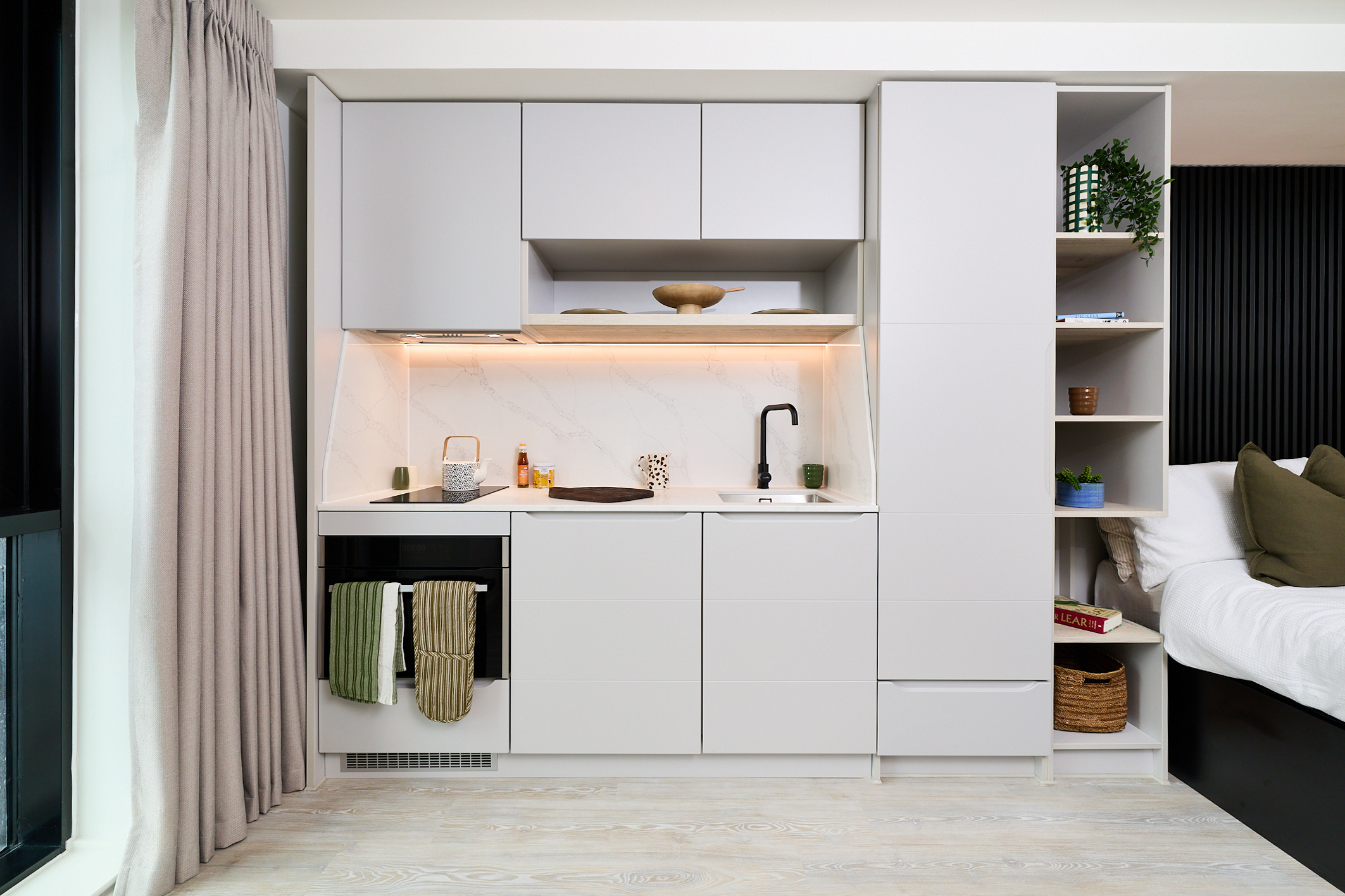 Modern compact kitchen with light gray cabinets, marble backsplash, open shelving, and under-cabinet lighting—perfectly suited for stylish urban living on Gough Street. Towels hang from the oven handle, and decor items are placed on the shelves. A bed is partially visible on the right.