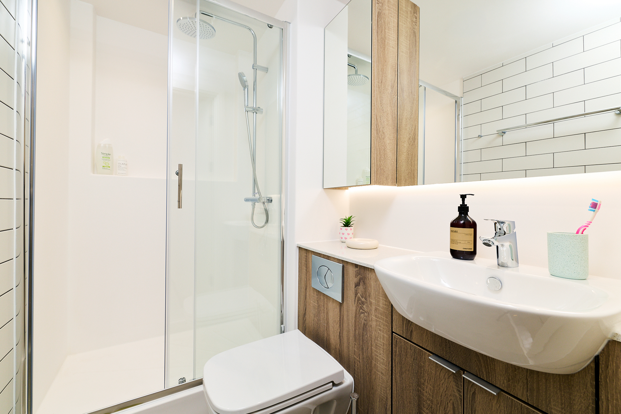 Modern Gough Street bathroom with a glass shower, wood cabinetry, white sink, soap dispenser, toothbrush holder, and wall tiles. Bright lighting and a clean, minimalist design.