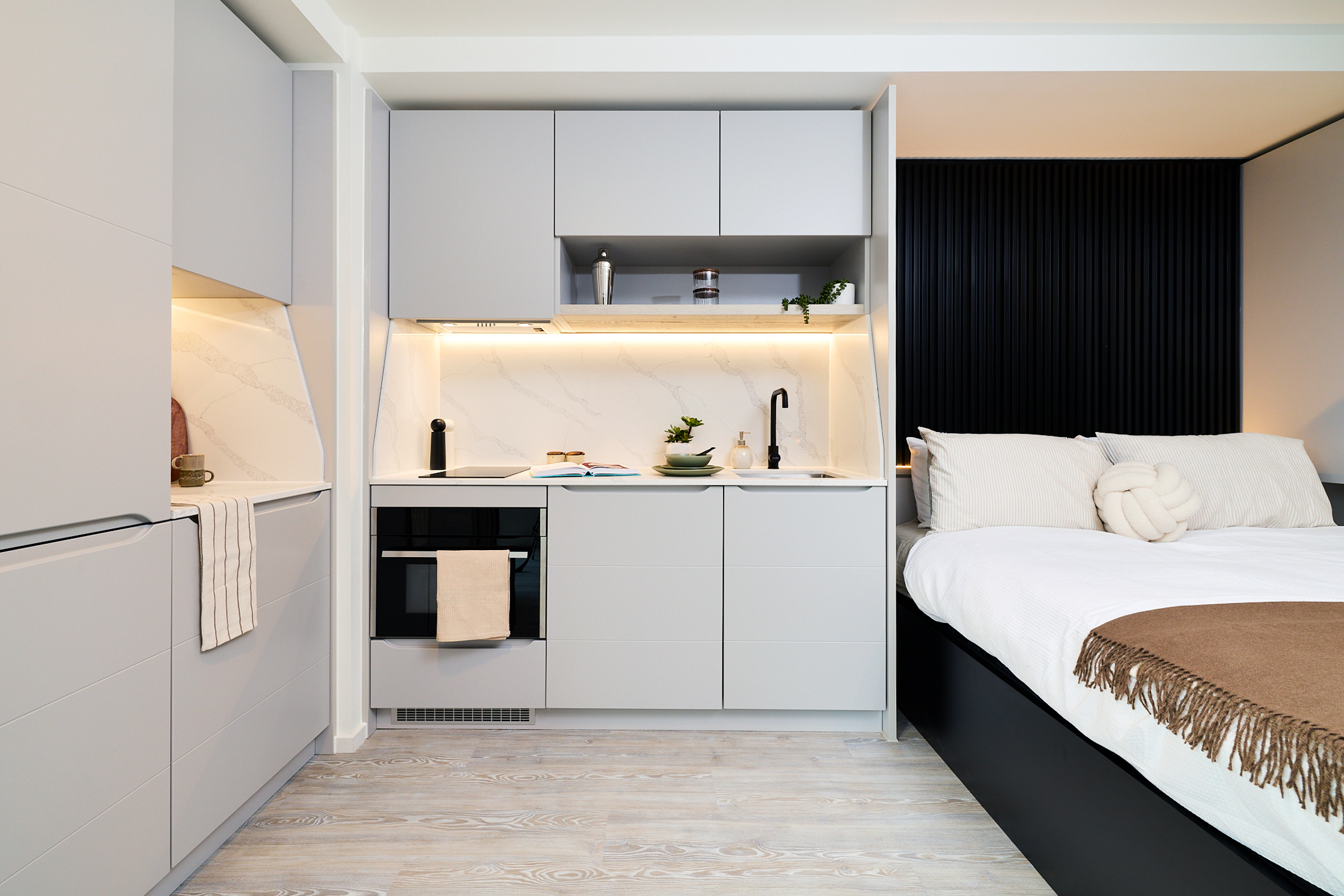 Modern studio apartment on Gough Street with a compact kitchen featuring light gray cabinets and built-in appliances, adjacent to a neatly made bed with white bedding and neutral-toned accents.