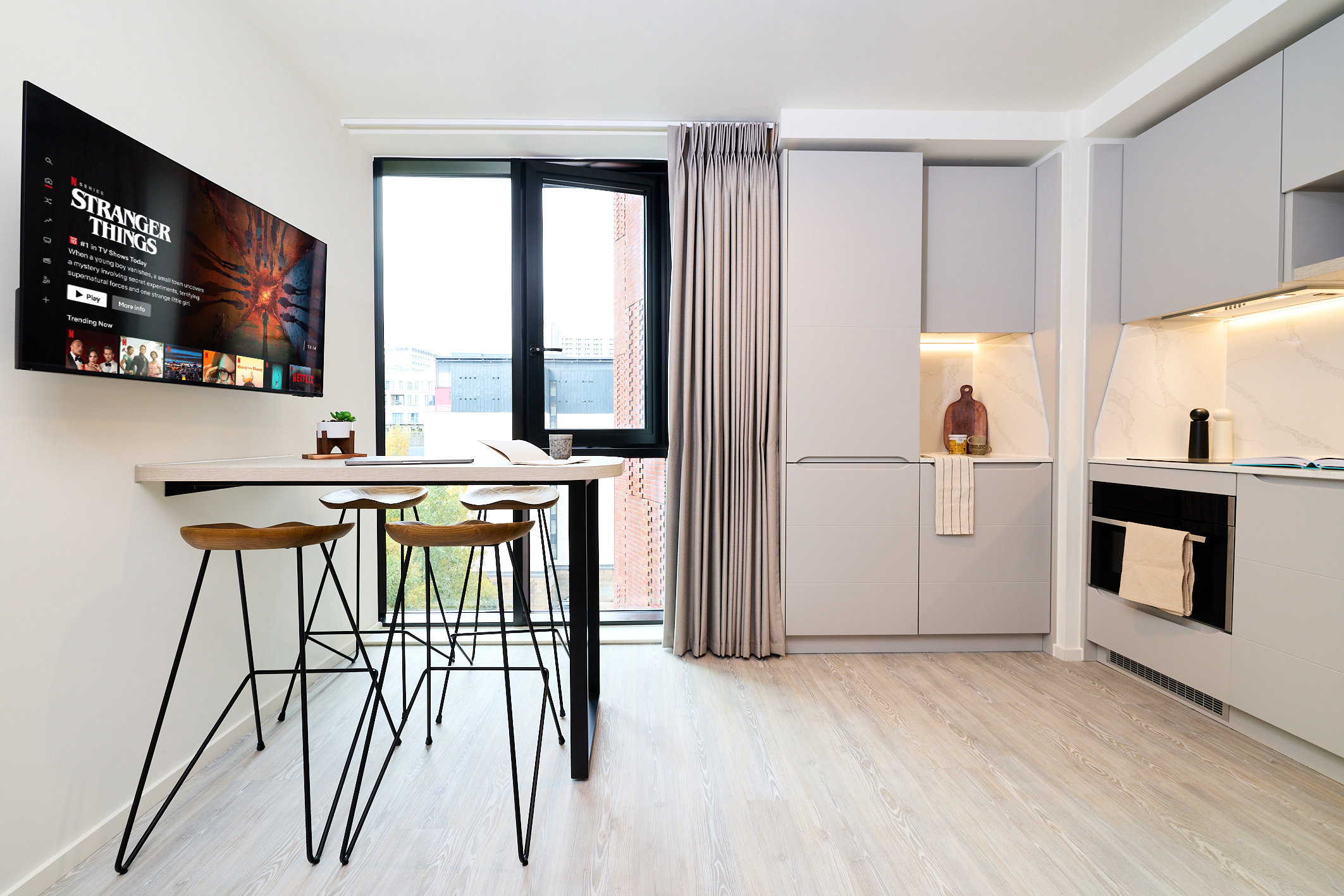 Modern kitchen with light gray cabinets, a built-in oven, and a high table with three bar stools, located along Gough Street. The space features a large window with curtains and a TV on the wall displaying Netflix’s Stranger Things.