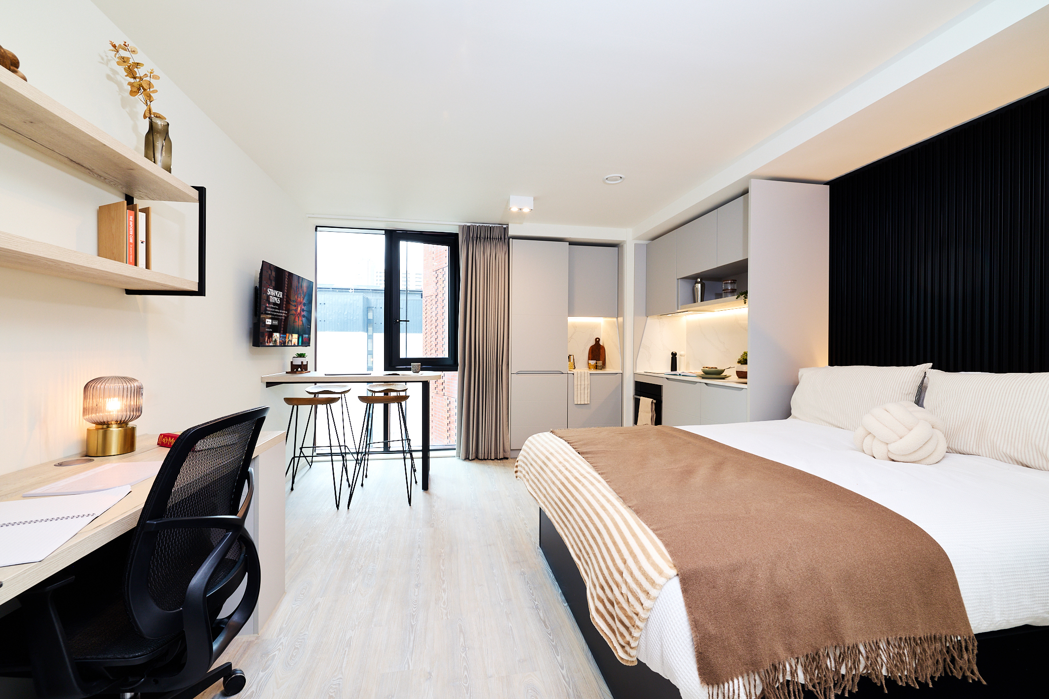 A modern studio apartment on Gough Street features a bed with neutral bedding, a kitchenette, a desk with a chair, wall shelves, a TV, and a window with two bar stools by a counter. The decor is minimal and stylish.