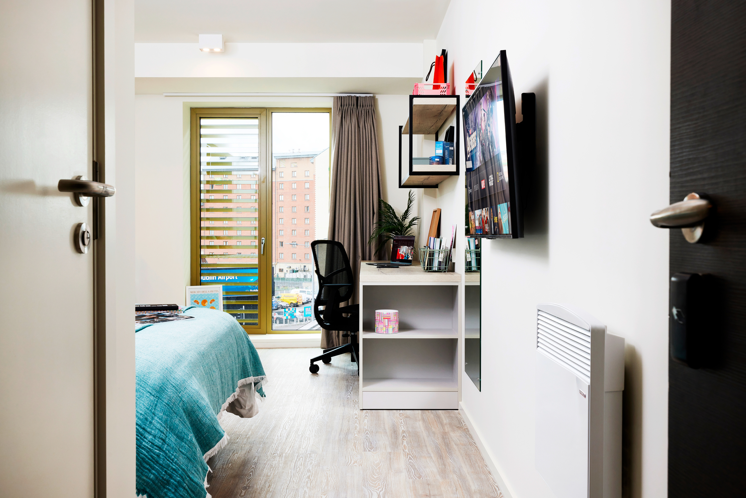 Modern student bedroom on Gough Street with a neatly made bed, desk, office chair, wall-mounted shelves, TV, and large window letting in natural light. Urban buildings are visible outside. The room features light-colored walls and flooring.