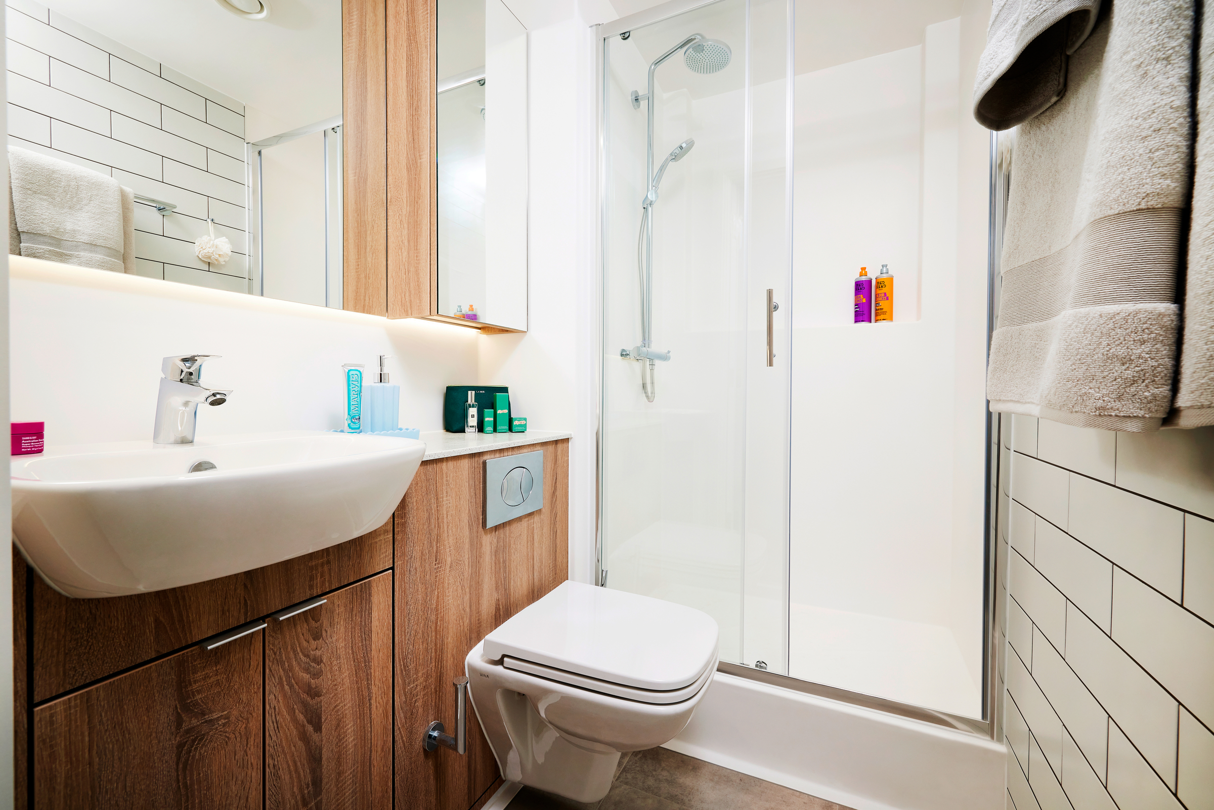 Modern Gough Street bathroom with a white sink and faucet, wall-mounted toilet, wooden cabinetry, and a glass-enclosed shower with toiletries. White tiled walls and neatly folded towels hang on the right.