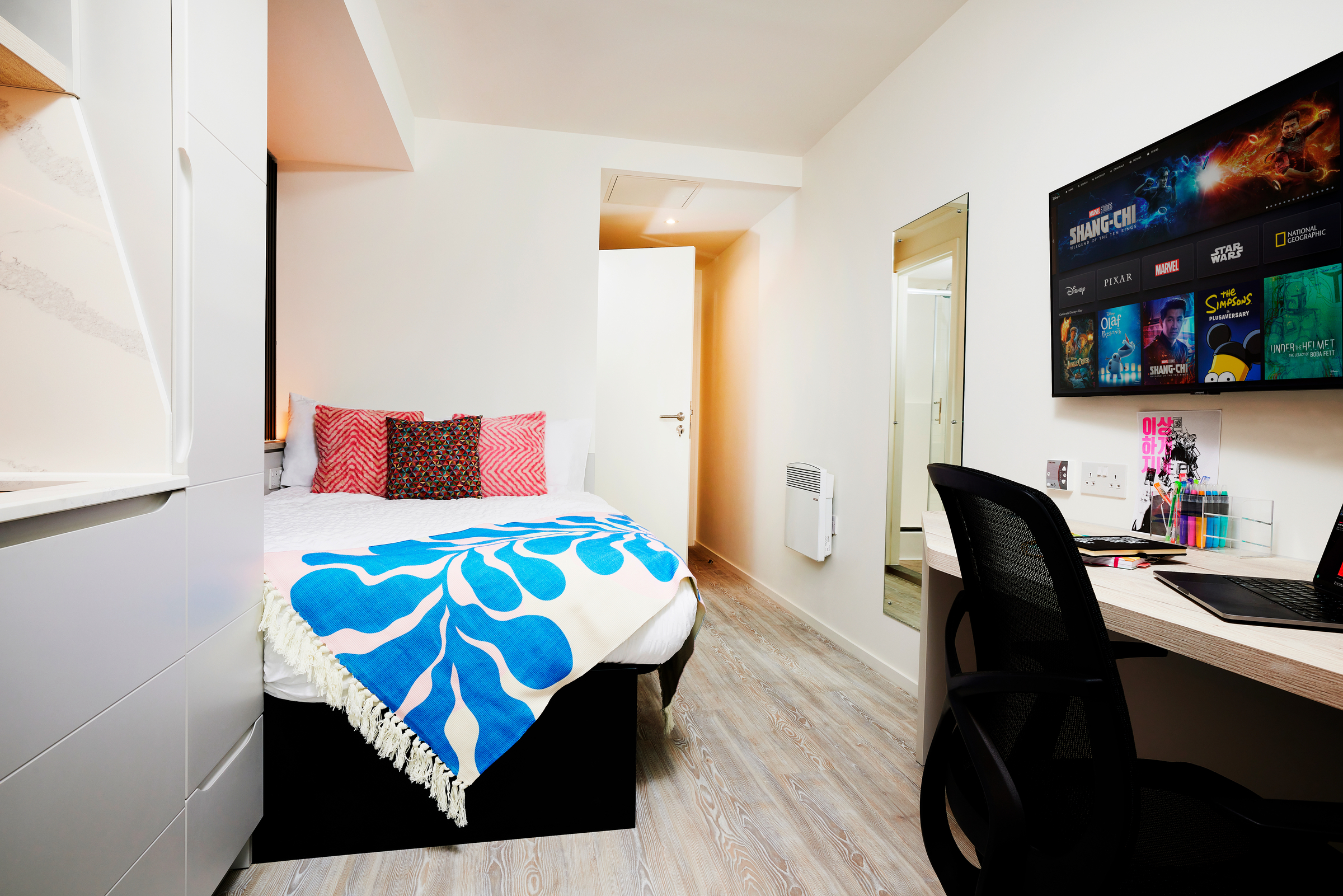 A modern, tidy bedroom on Gough Street with a bed featuring red pillows and a blue-and-white blanket. There’s a desk with chair, monitor displaying movie posters, and a wall mirror. Light wood flooring completes the look.