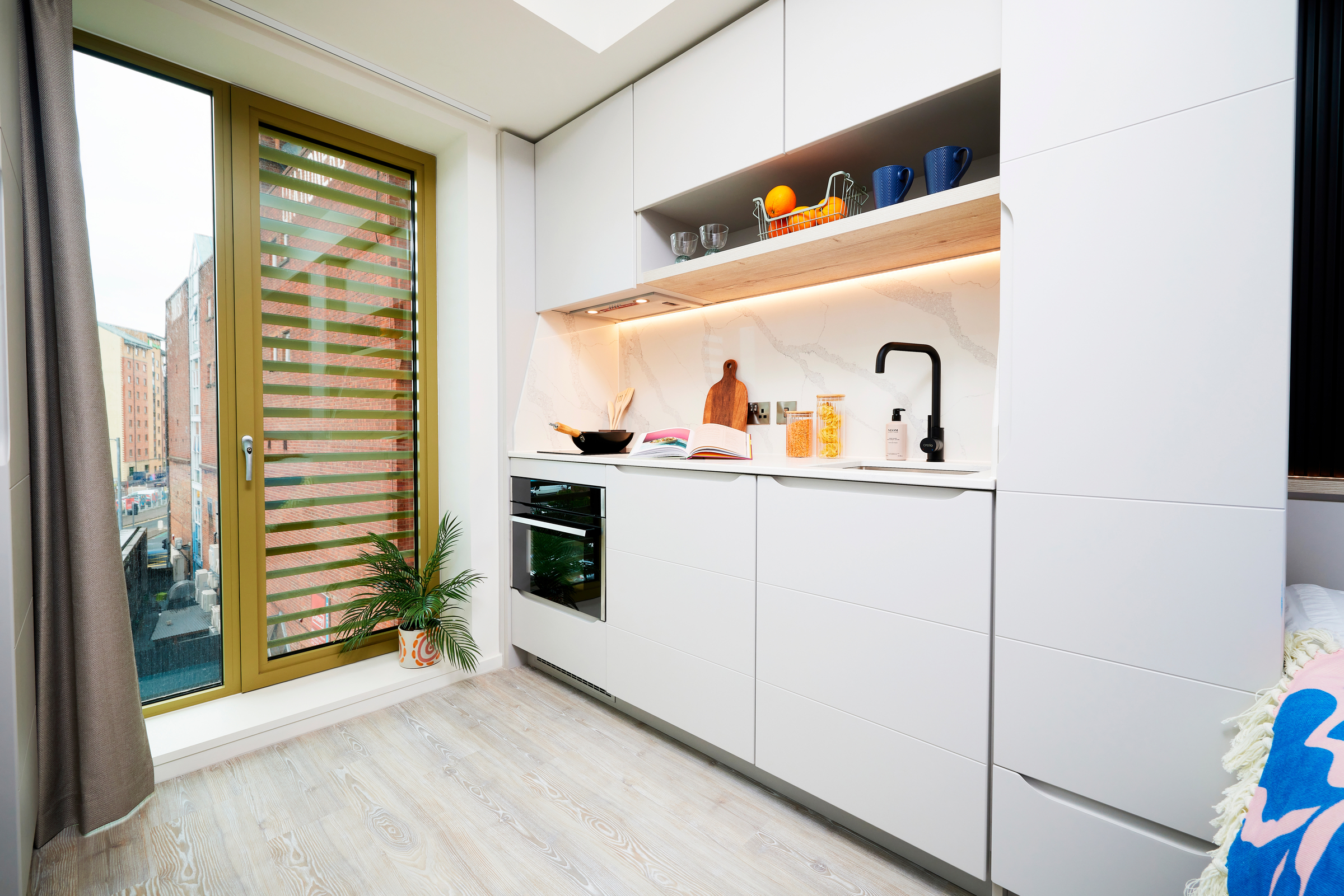 Modern, compact kitchen on Gough Street with white cabinets, built-in oven, black faucet, and marble backsplash. Open shelves display mugs and fruit. A large window and potted plant add light and greenery to the bright space.