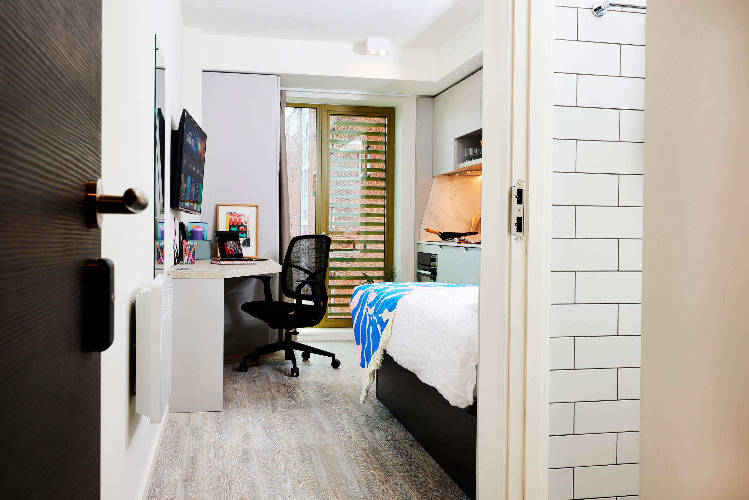 Modern Gough Street bedroom with a desk, office chair, wall-mounted TV, and bed with blue blanket. Features include wooden flooring, a window with blinds, and a partially visible white-tiled bathroom on the right.