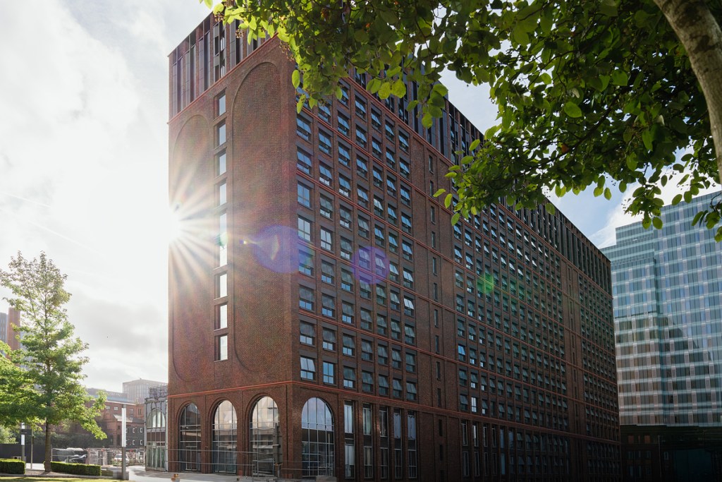 A modern red-brick building with large arched windows at the base and many rectangular windows above, ideal for students moving to Manchester, with sunlight shining from behind and green trees in the foreground.