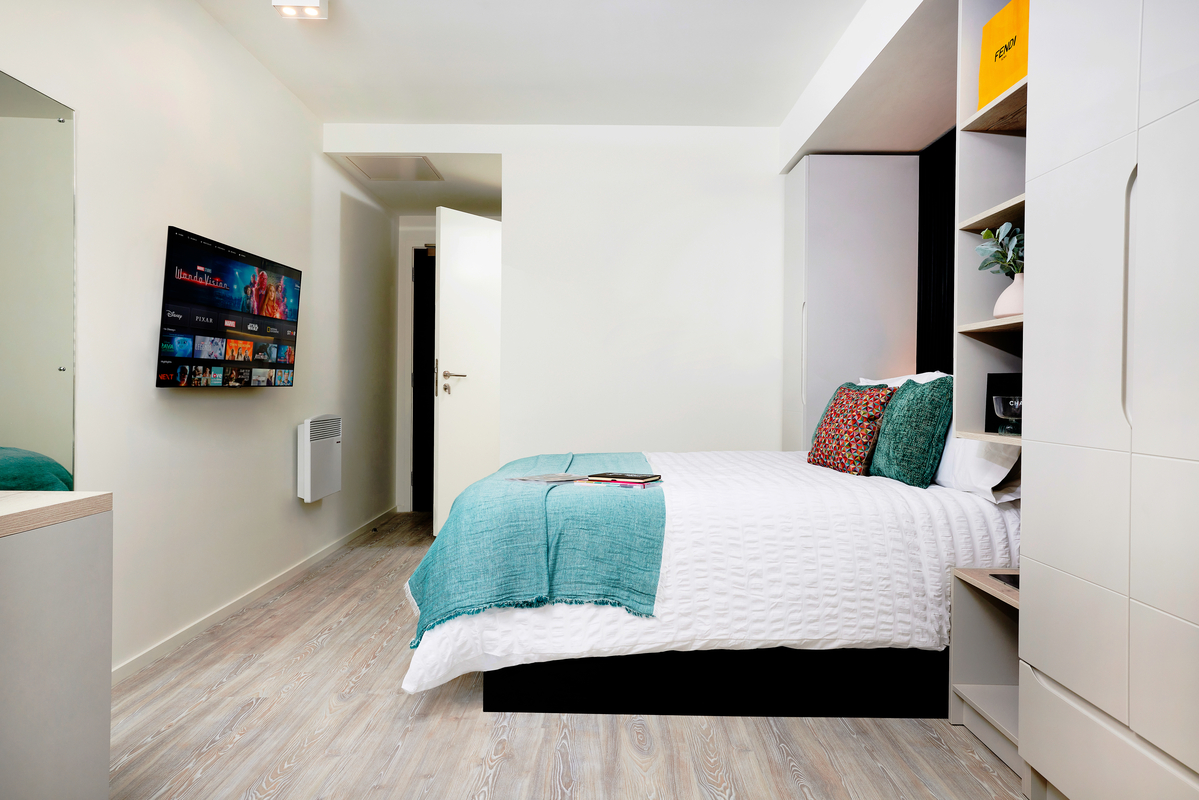 Modern bedroom with a neatly made bed featuring teal accents, a wall-mounted TV displaying streaming apps, white walls, wooden floor, open door, and built-in storage units with decorative items.