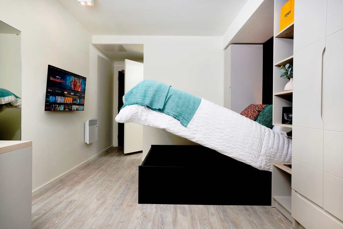 A modern, compact bedroom with a lift-up bed revealing storage space underneath, a wall-mounted TV displaying streaming services, light wood flooring, and built-in shelves and closet for organization.