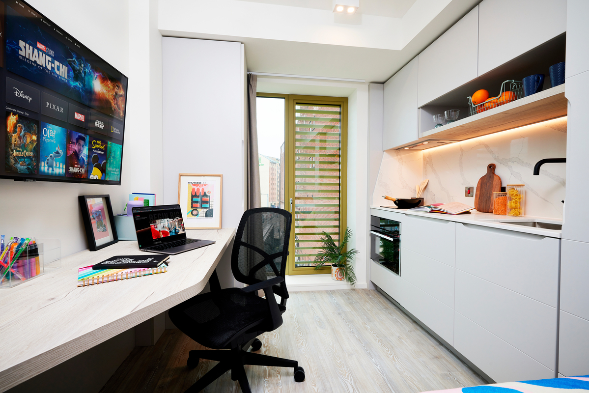 Compact, modern studio apartment with a wall-mounted TV, corner desk and chair, laptop, kitchen area with white cabinets, sink, oven, and shelves, potted plant by a door, and colorful decor.