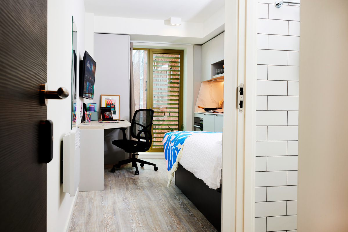 A tidy, modern dorm room with a bed, a desk and chair, wall-mounted TV, shelves, and a window with wooden blinds. The room has light wood flooring and a partially visible tiled bathroom.