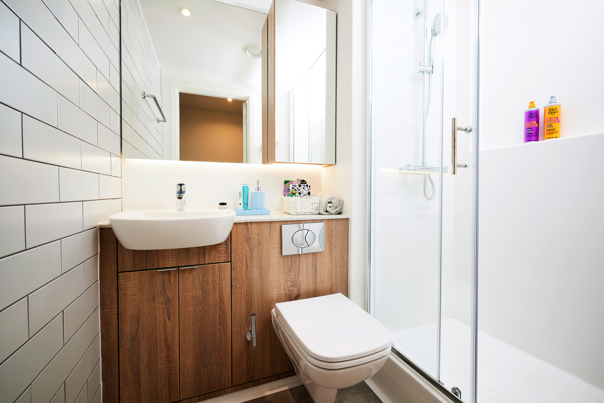 A modern bathroom with white subway tiles, a wood vanity with a sink, a wall-mounted toilet, and a glass shower. Toiletries are on the sink and in the shower, and a large mirror reflects the room.