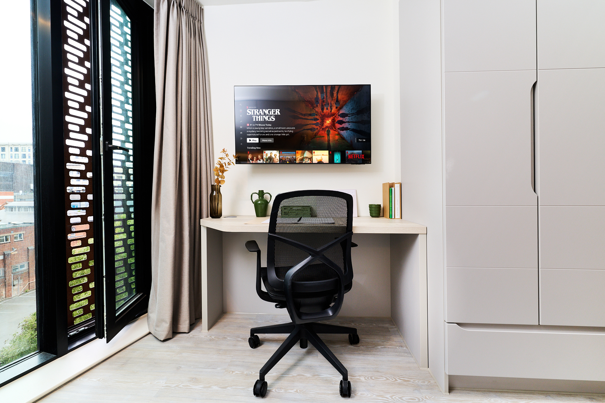 A modern Gough Street home office with a desk, chair, laptop, plant, and books. A TV on the wall displays the show “Stranger Things.” Large windows with curtains let in natural light.