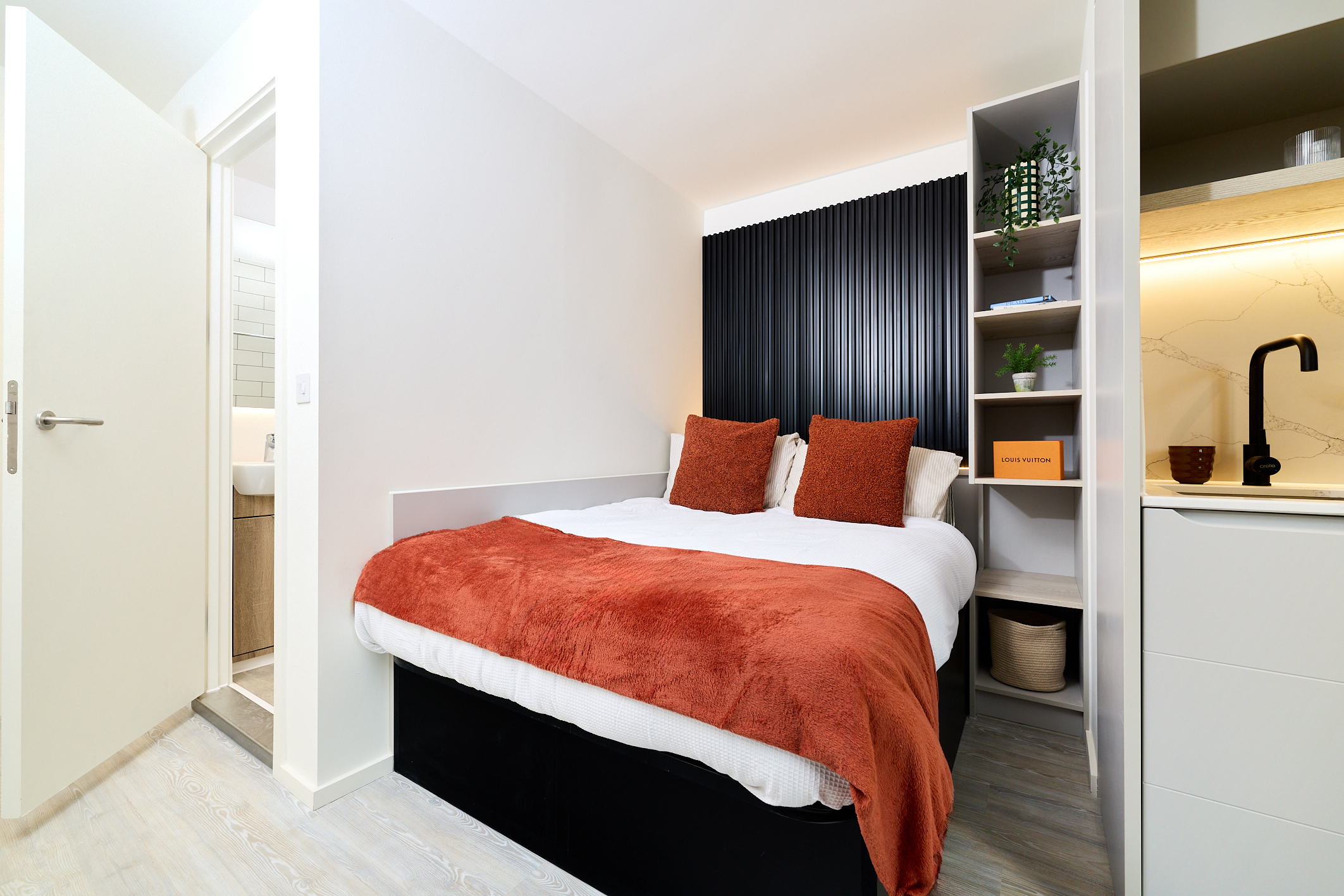 A modern, compact bedroom on Gough Street features a double bed with rust-colored pillows and blanket, built-in shelves, a kitchenette, and a door leading to a small bathroom.