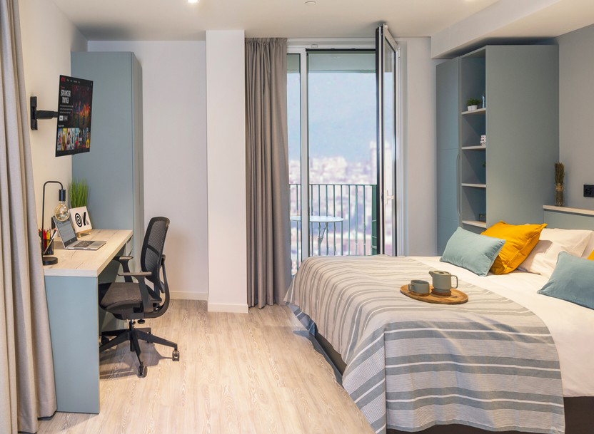A modern, tidy bedroom with a double bed, striped bedding, and yellow pillows. A desk with a chair, lamp, and computer sits by a window. Curtains and a door open to a balcony with a city and mountain view.