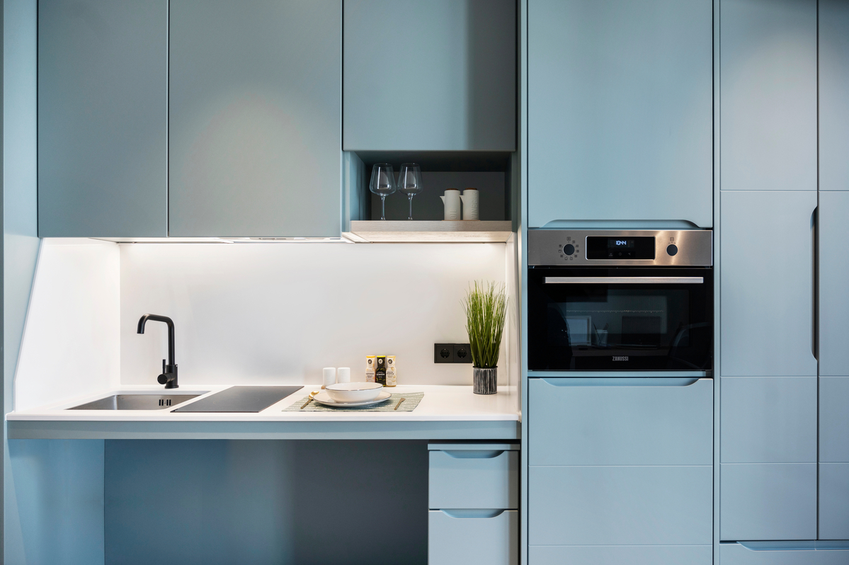 Modern kitchen with sleek light blue cabinets, built-in oven, black faucet, a sink, induction cooktop, potted plant, and open shelf holding glasses and cups. Minimalist design with clean lines and bright lighting.
