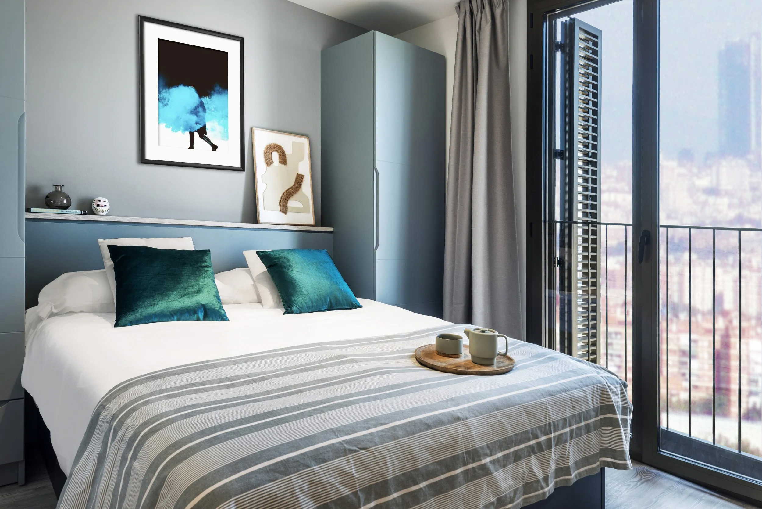 A modern bedroom with a double bed, teal pillows, striped blanket, framed abstract art on the wall, and a glass door opening to a city view. A tray with a teapot and cups sits on the bed.