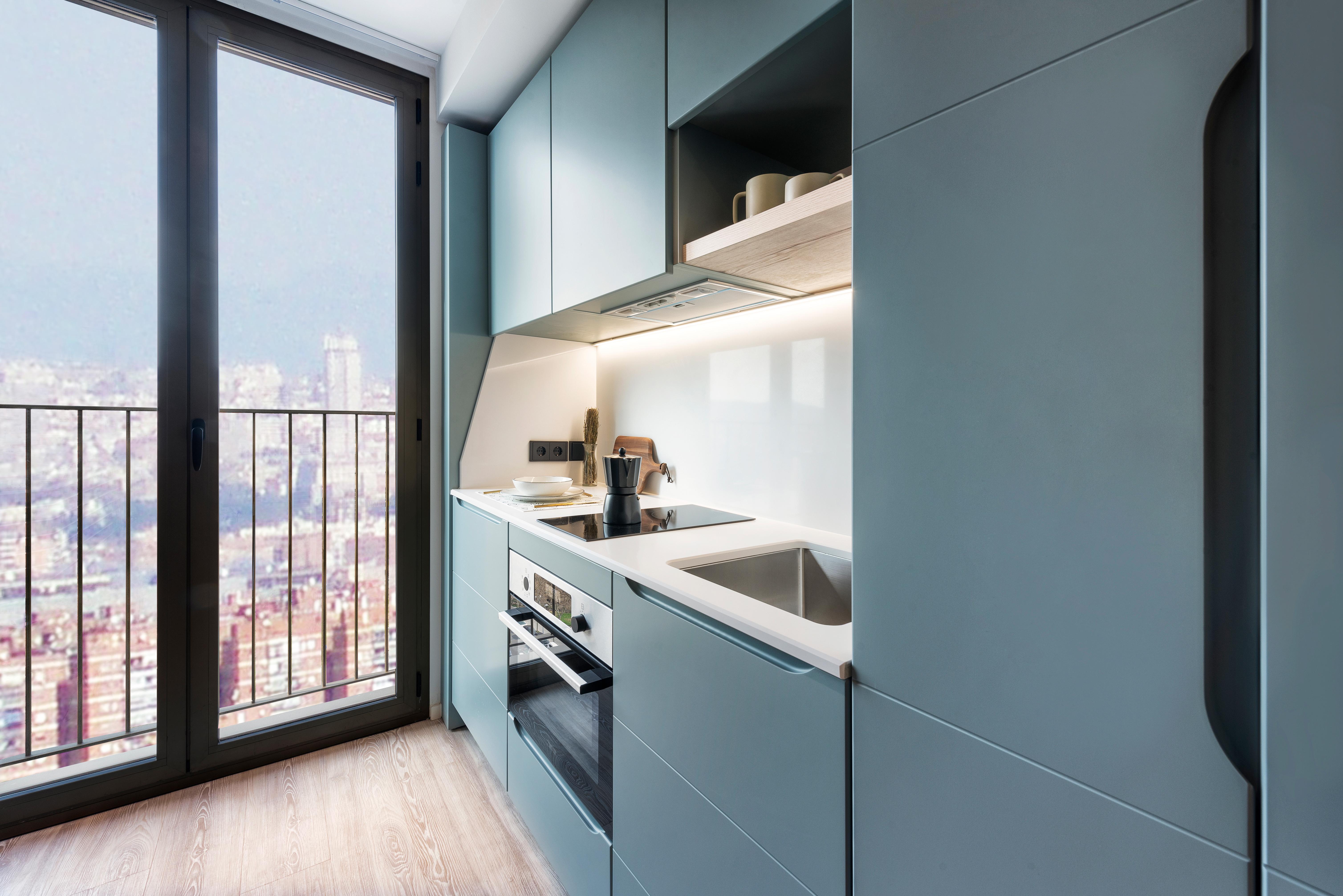 Modern compact kitchen with light blue cabinets, built-in appliances, and a white countertop. Large window with city view lets in natural light. Cookware and dishes are neatly arranged on the shelves.