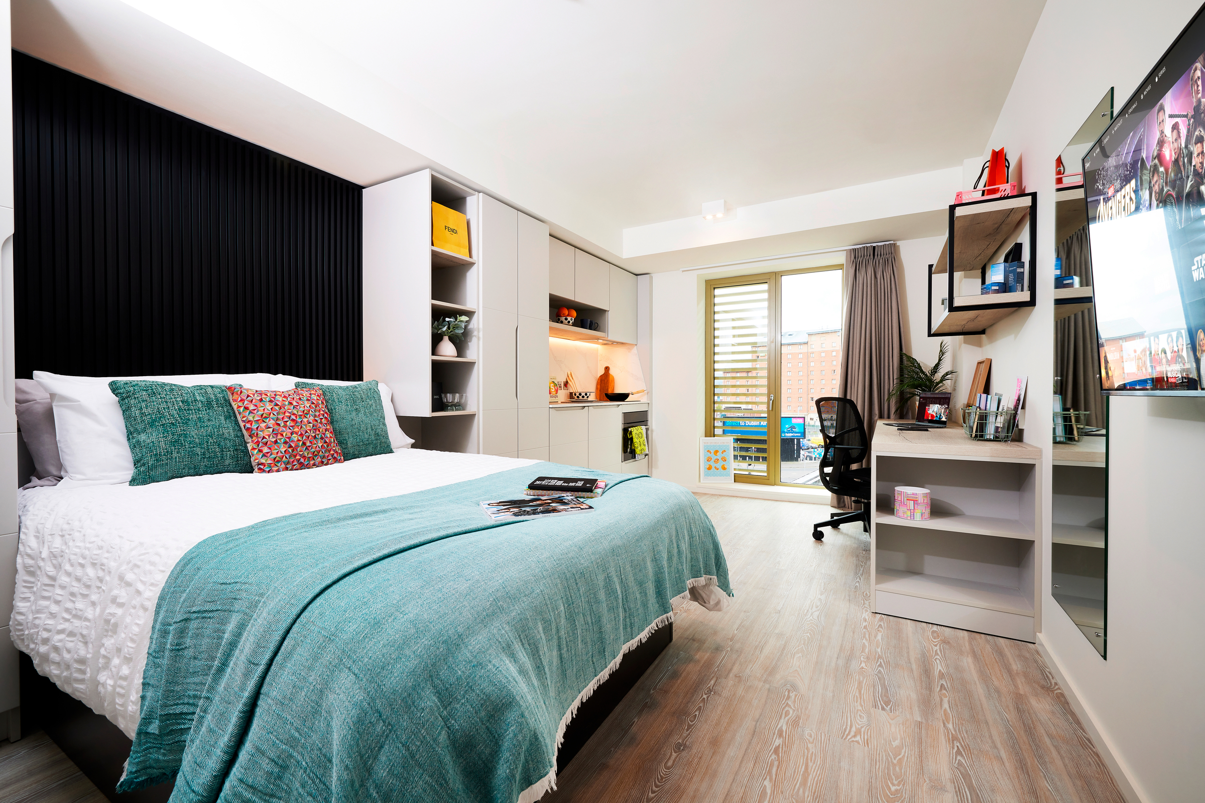 Modern bedroom in New Waverley with a large bed, teal blanket, colorful pillows, wall shelves, a desk and chair, and a TV mounted on the wall. Glass doors open to a small balcony with outdoor seating.