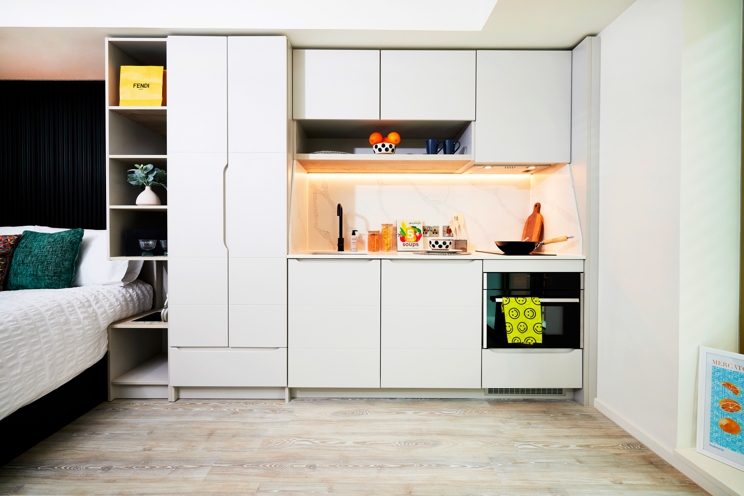 Modern, compact kitchen at New Waverley with white cabinets, open shelves, stove, sink, and subtle under-cabinet lighting. A bed is partially visible on the left; colorful decor and cookware add warmth to the clean design.