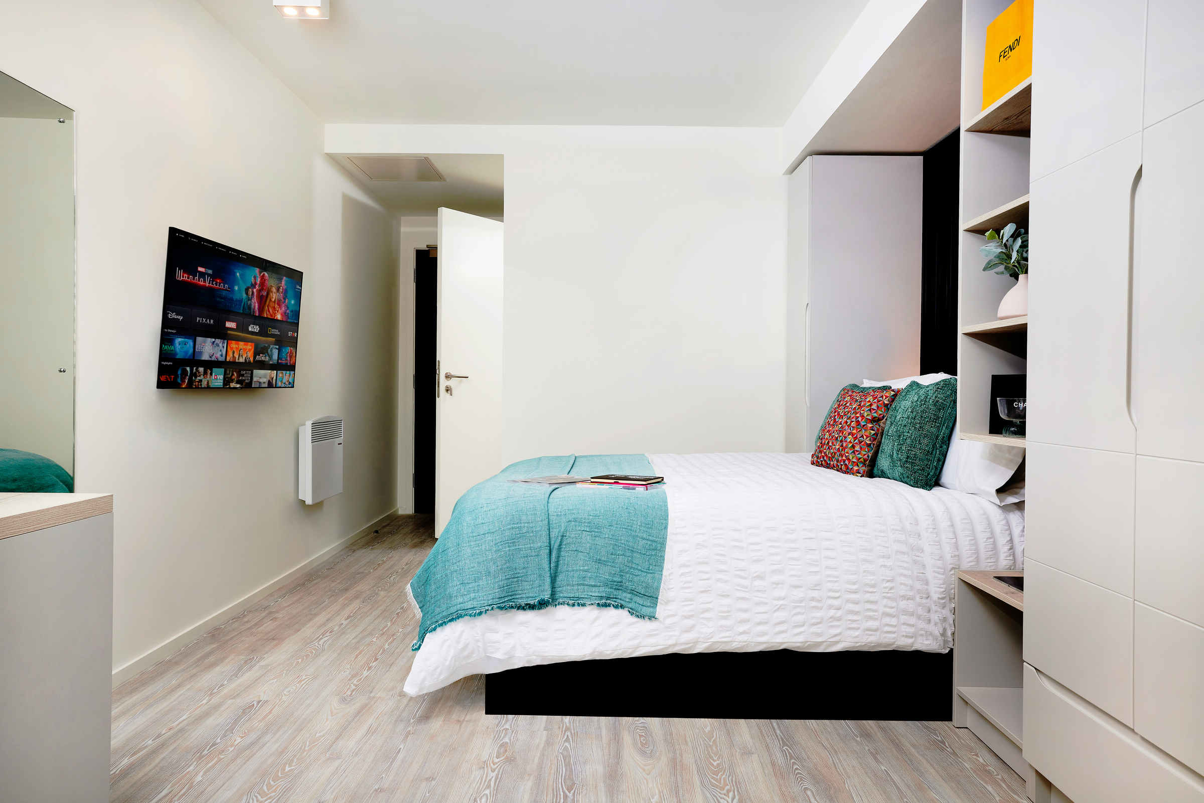 A modern, tidy bedroom at New Waverley with a white bed, teal throw and pillows, a wall-mounted TV displaying streaming apps, open shelving with decor, light wood flooring, and a partially open door leading to another room.