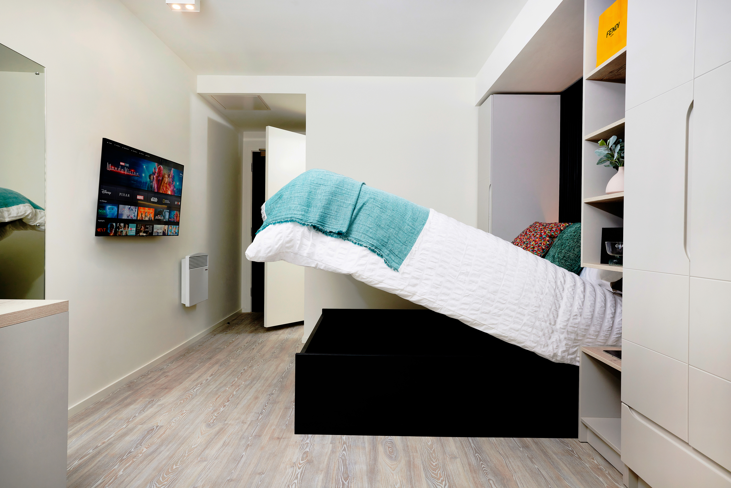 A modern New Waverley bedroom with light wood flooring, a mounted TV, and a bed lifted to reveal storage underneath. The white walls are complemented by built-in shelves and cabinets beside the bed.
