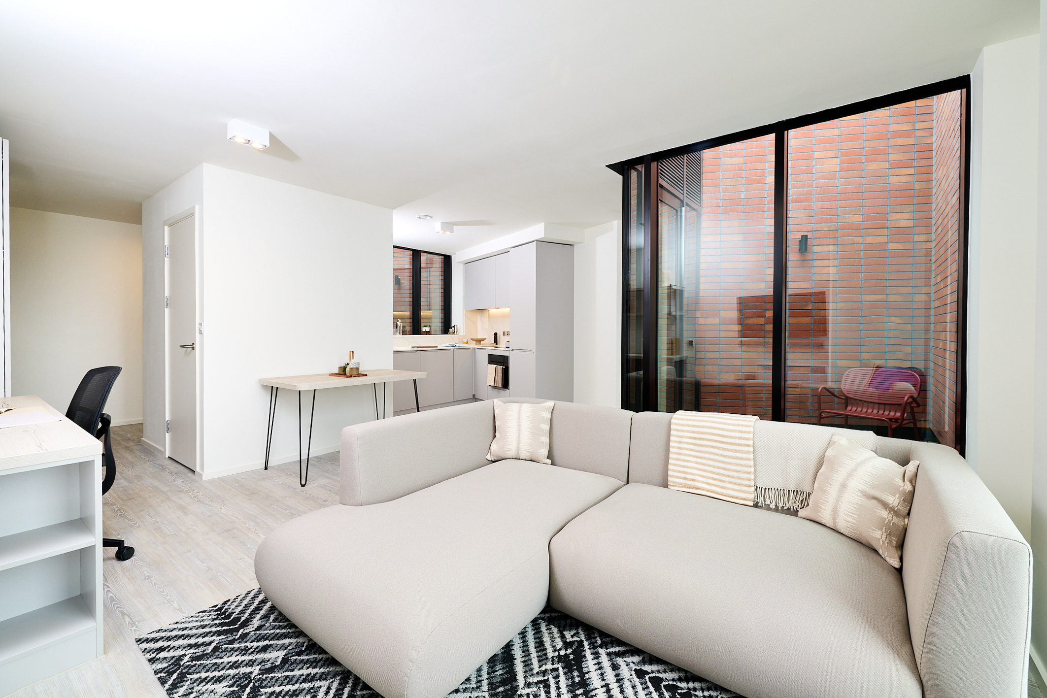 Modern apartment living area with a light gray sectional sofa, patterned rug, wall desk with chair, dining table, and open kitchen. Large windows reveal a small patio with a chair and brick walls, offering elegant urban views that capture the vibrant atmosphere of Gough Street.