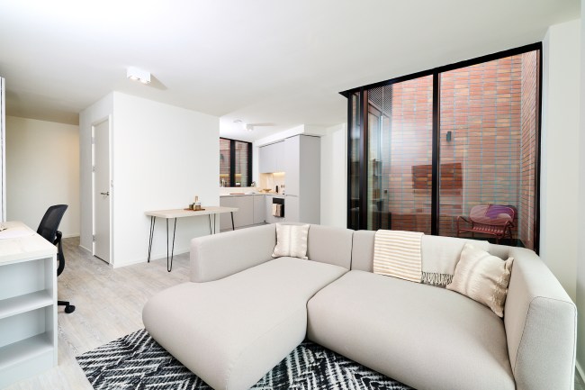 Modern apartment living area with a light gray sectional sofa, patterned rug, wall desk with chair, dining table, and open kitchen. Large windows reveal a small patio with a chair and brick walls, offering elegant urban views that capture the vibrant atmosphere of Gough Street.