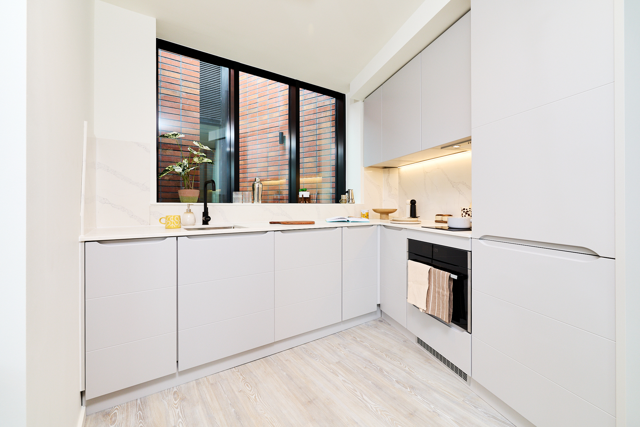 Modern white kitchen on Gough Street with minimalist cabinets, built-in oven, and a large window showing a brick courtyard. Light wood flooring and clean countertops with simple decor create a bright, airy atmosphere.