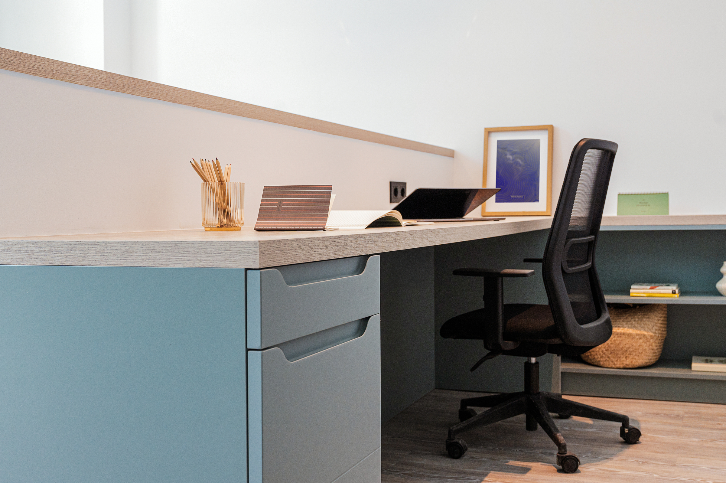 Modern office workspace with a black desk chair, a blue desk, pencils in a clear holder, a notebook, framed artwork, a basket, and a green book on a shelf under the desk. Bright, minimalist decor.