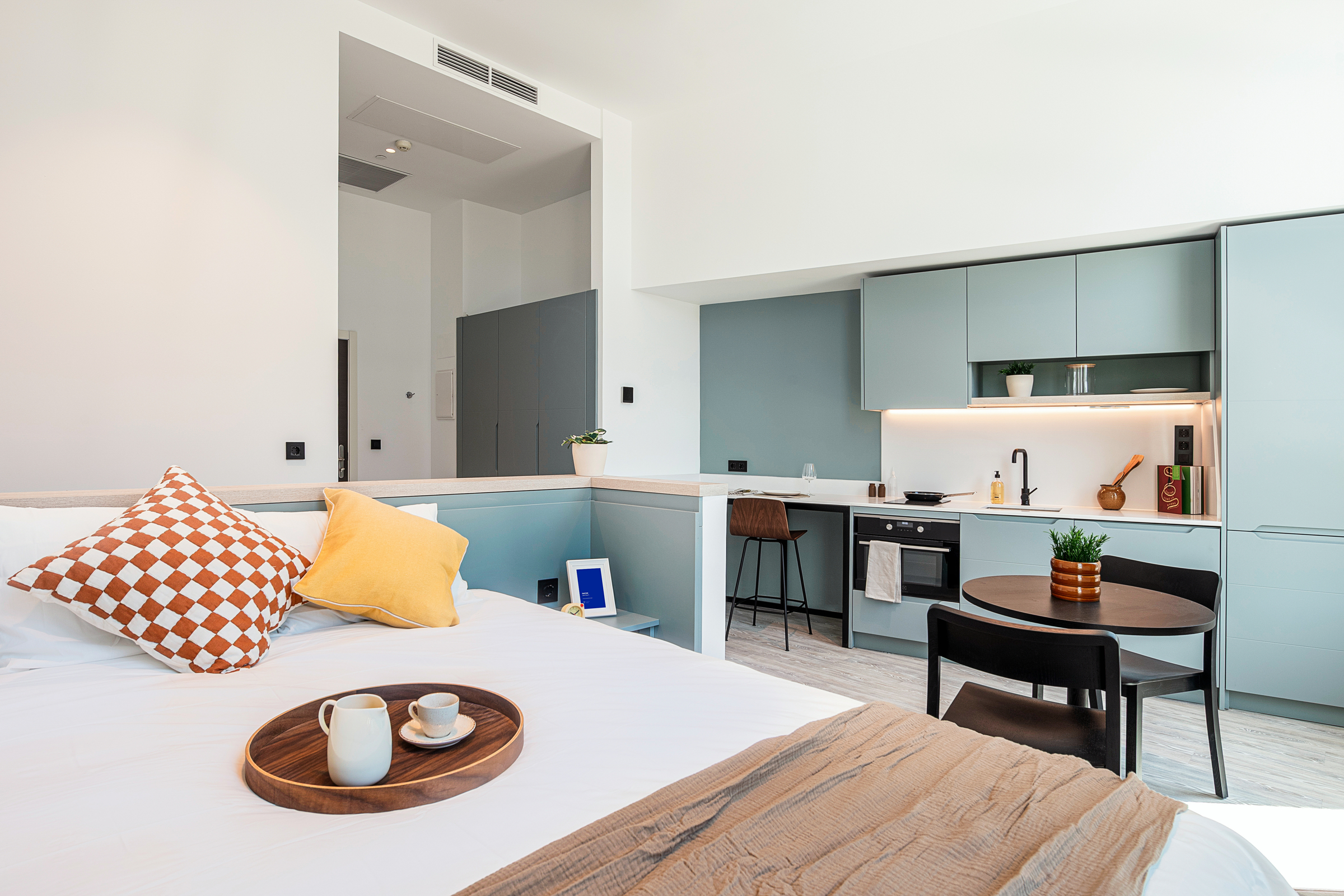 Bright studio apartment with a bed, a tray with coffee cups, a modern kitchen with blue cabinets, built-in appliances, a small dining table, and minimalist decor.