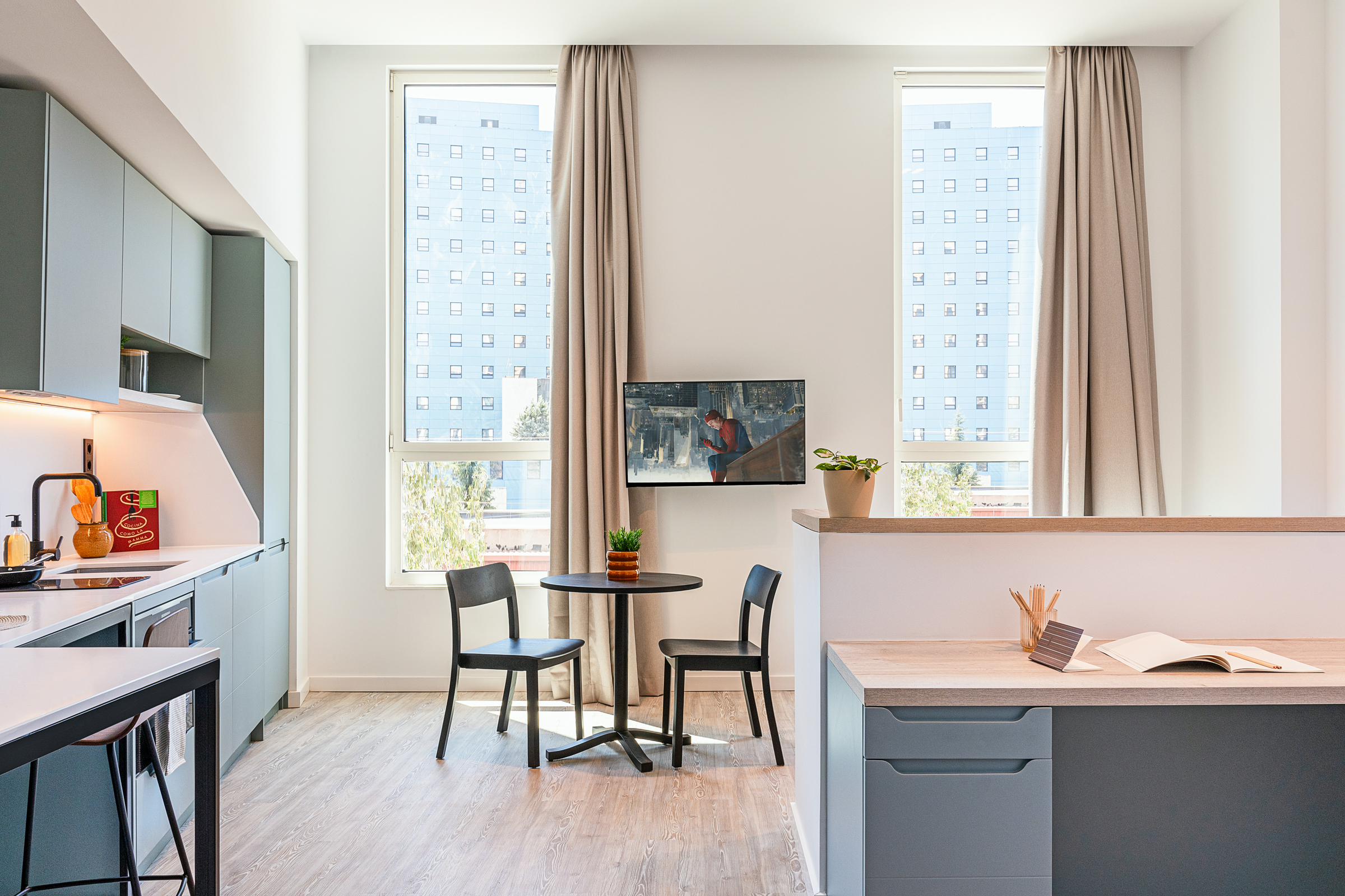 Modern kitchen and dining area with two black chairs and a round table near tall windows, beige curtains, TV on wall, and adjacent workspace with open notebook and pencils.