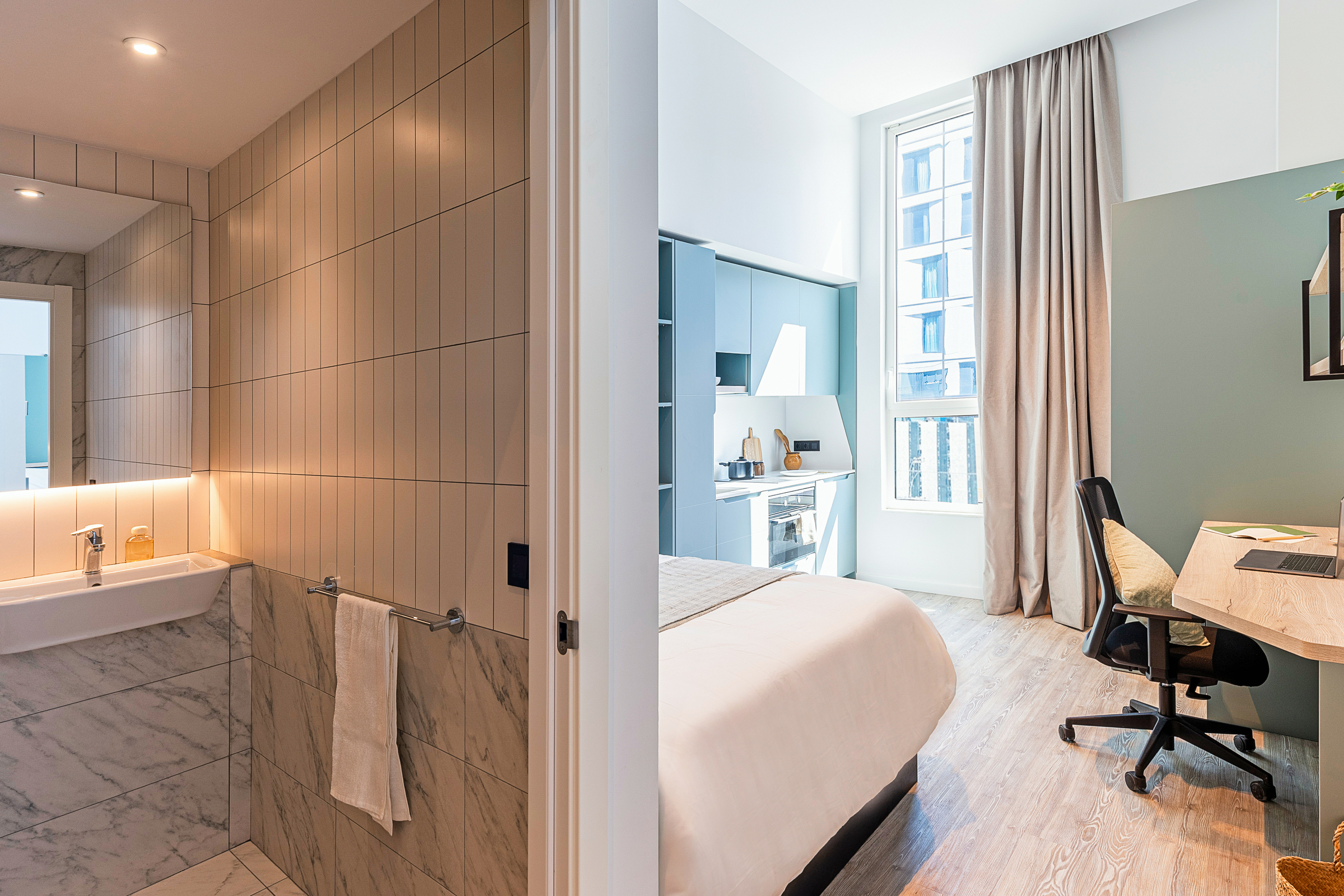 Modern studio apartment with a bathroom on the left, featuring a sink and tiled walls, and a bedroom with a neatly made bed, desk, chair, and large window on the right.