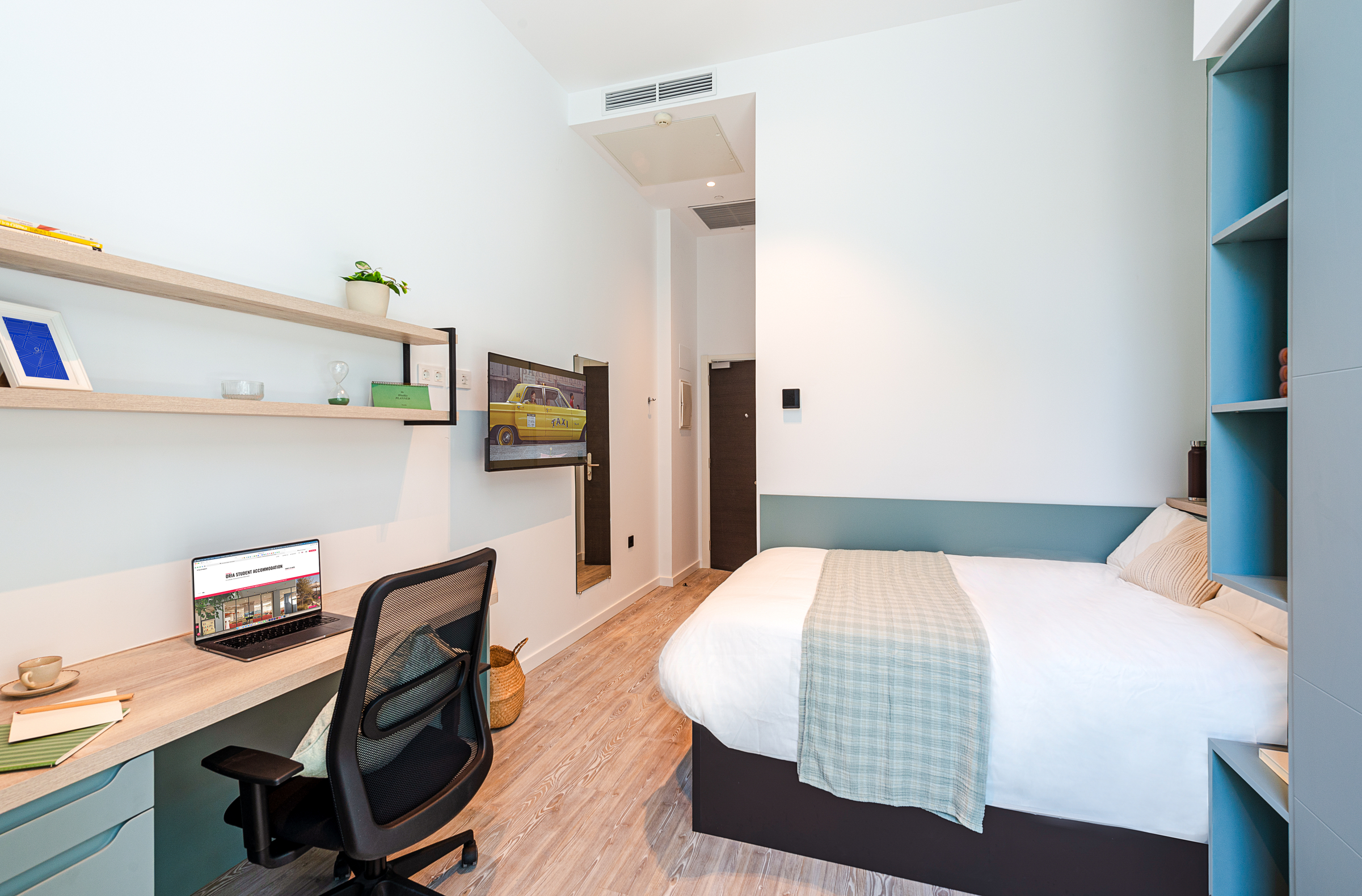 A modern, tidy bedroom with a double bed, shelves, a desk with a laptop, an office chair, and a wall-mounted TV. Light wood flooring and blue accents create a cozy, minimalist atmosphere.
