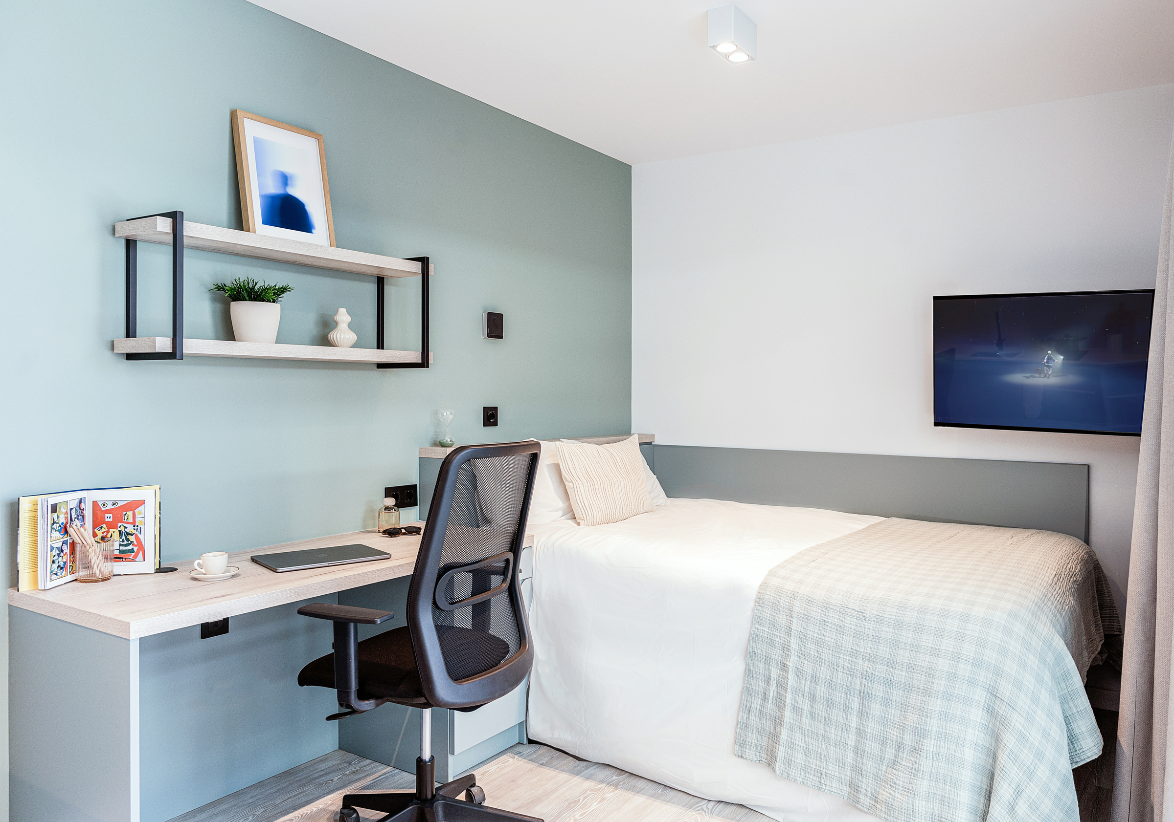 Modern, tidy bedroom with a made bed, checked beige blanket, desk and office chair, wall shelves with decor, a cup and books on the desk, and a wall-mounted TV showing a dark scene.