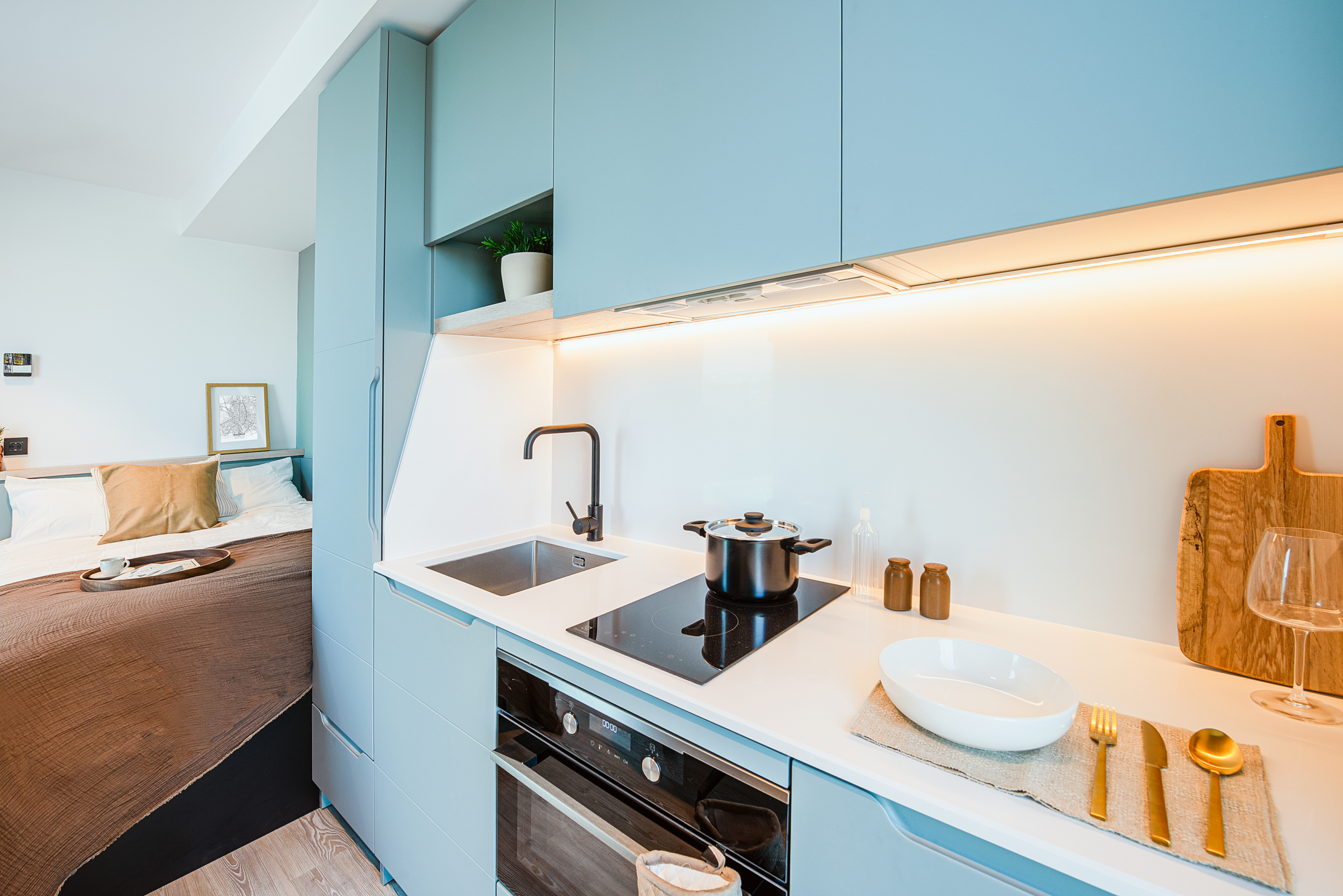 Modern studio kitchen with blue cabinets, induction cooktop, pot, sink, and neatly set tableware, adjacent to a cozy bed with brown bedding and framed art. Warm lighting brightens the space.