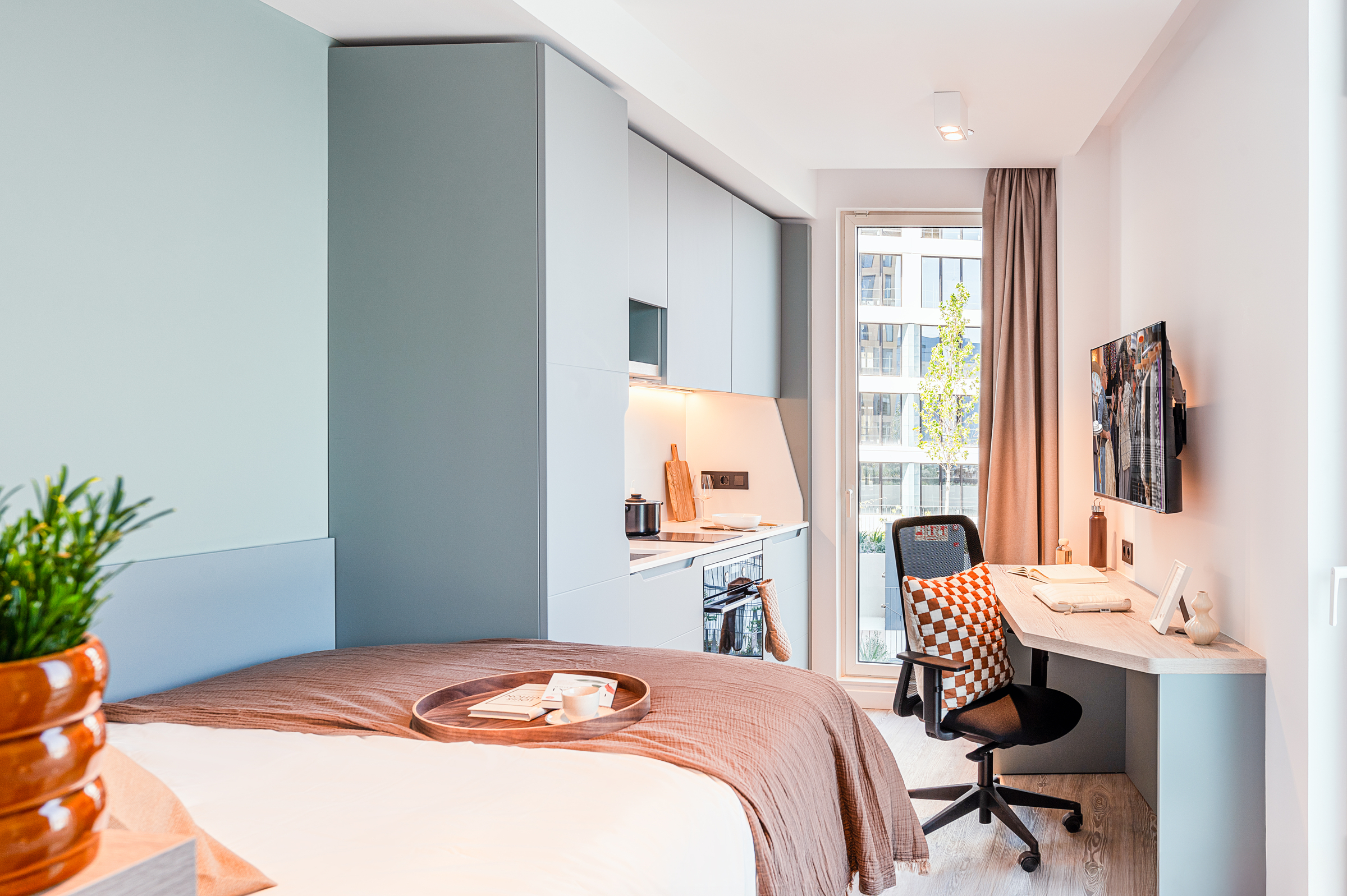 A modern studio apartment with a neatly made bed, kitchenette, desk with office chair, wall-mounted TV, large window, and potted plant, featuring soft neutral colors and natural light.