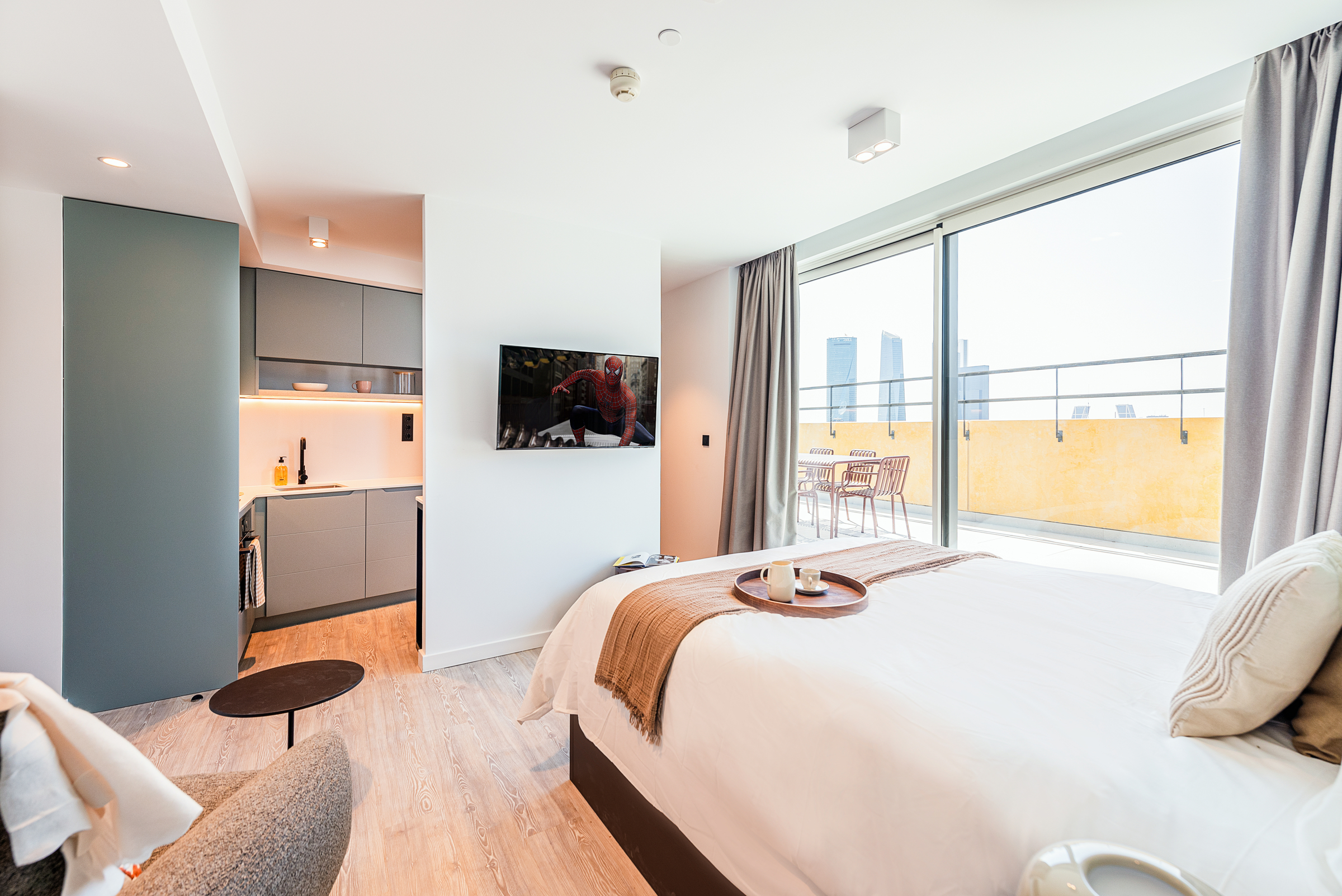 Modern, sunlit hotel room with a large bed, tray with coffee, wall-mounted TV, kitchenette, and floor-to-ceiling windows leading to a balcony overlooking city buildings.