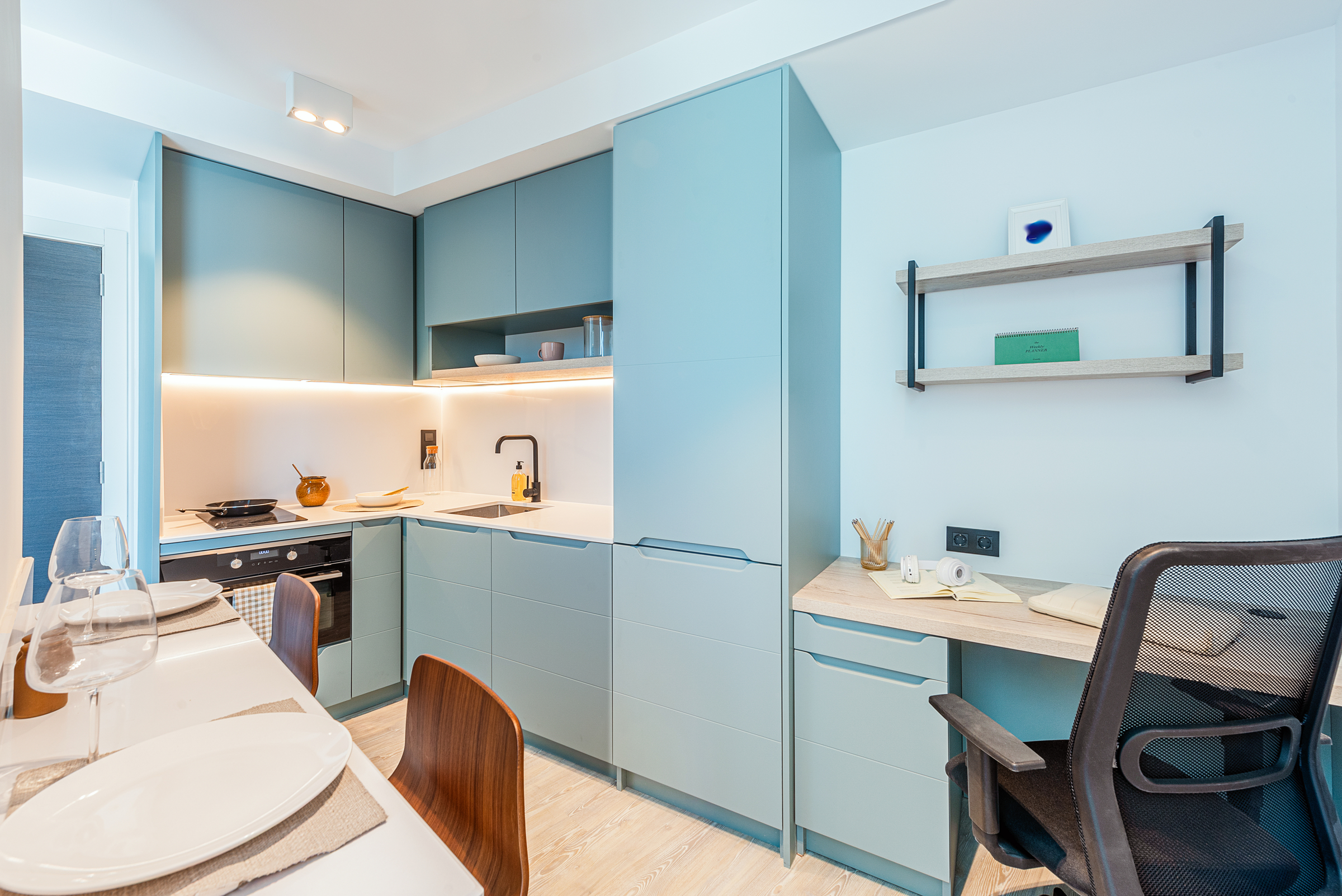 Modern kitchen with teal cabinets, built-in appliances, a small desk with a chair, open shelving, and a dining table set with plates and glasses. The space is bright and neatly organized.