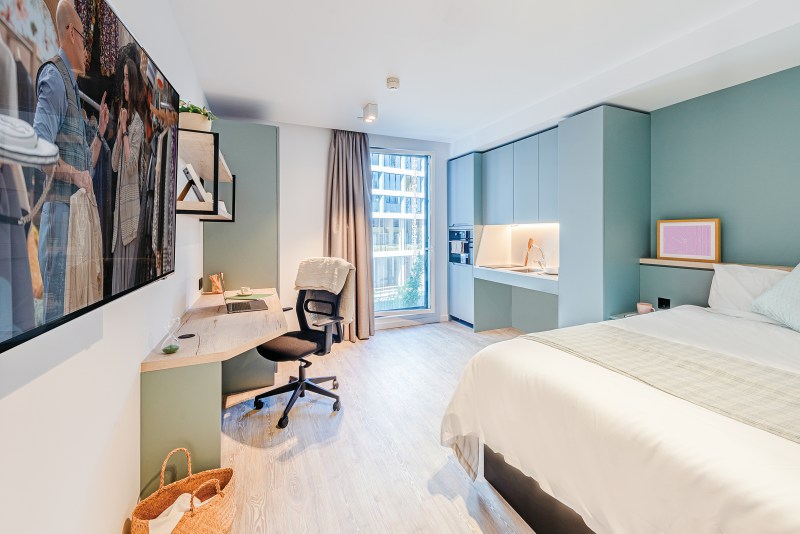A modern, bright bedroom with a large bed, wall-mounted TV, desk and chair, open shelving, a kitchenette area, and a large window with curtains letting in natural light.