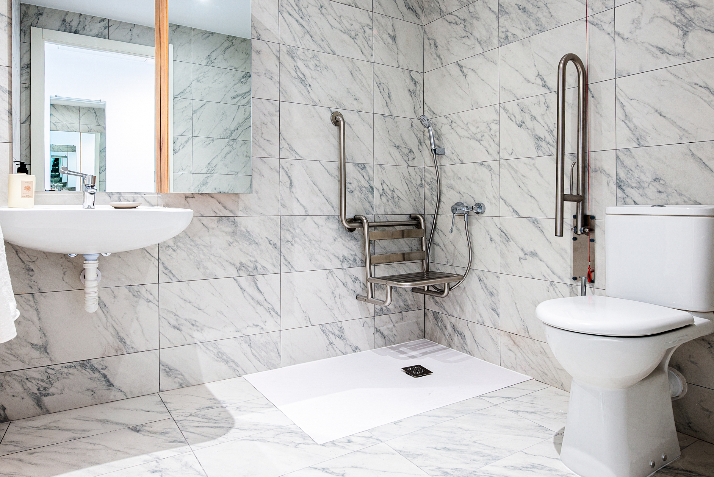Accessible bathroom with a wall-mounted sink, toilet, walk-in shower area, fold-down shower seat, and metal grab bars on the walls. All surfaces are tiled in white with gray marbling.