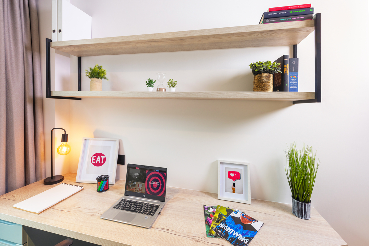 A tidy desk with a laptop, magazines, framed art, a notepad, lamp, and potted plants. Two shelves above hold more plants, books, and decor. The setup is bright and organized, ideal for studying or working.