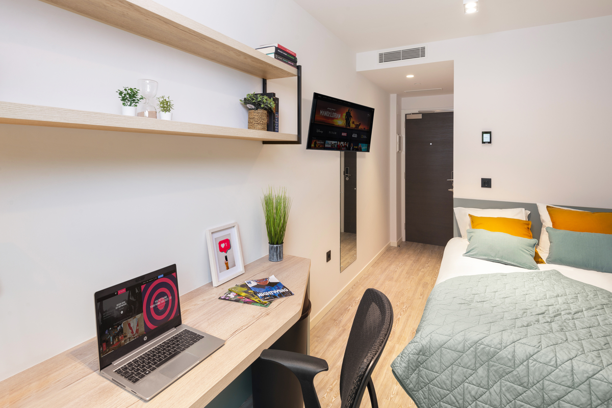 Modern student room with a bed, desk, laptop, chair, shelves with plants and books, a door at the back, a TV on the wall, and a light wood floor. The decor is minimalist with green and orange accents.