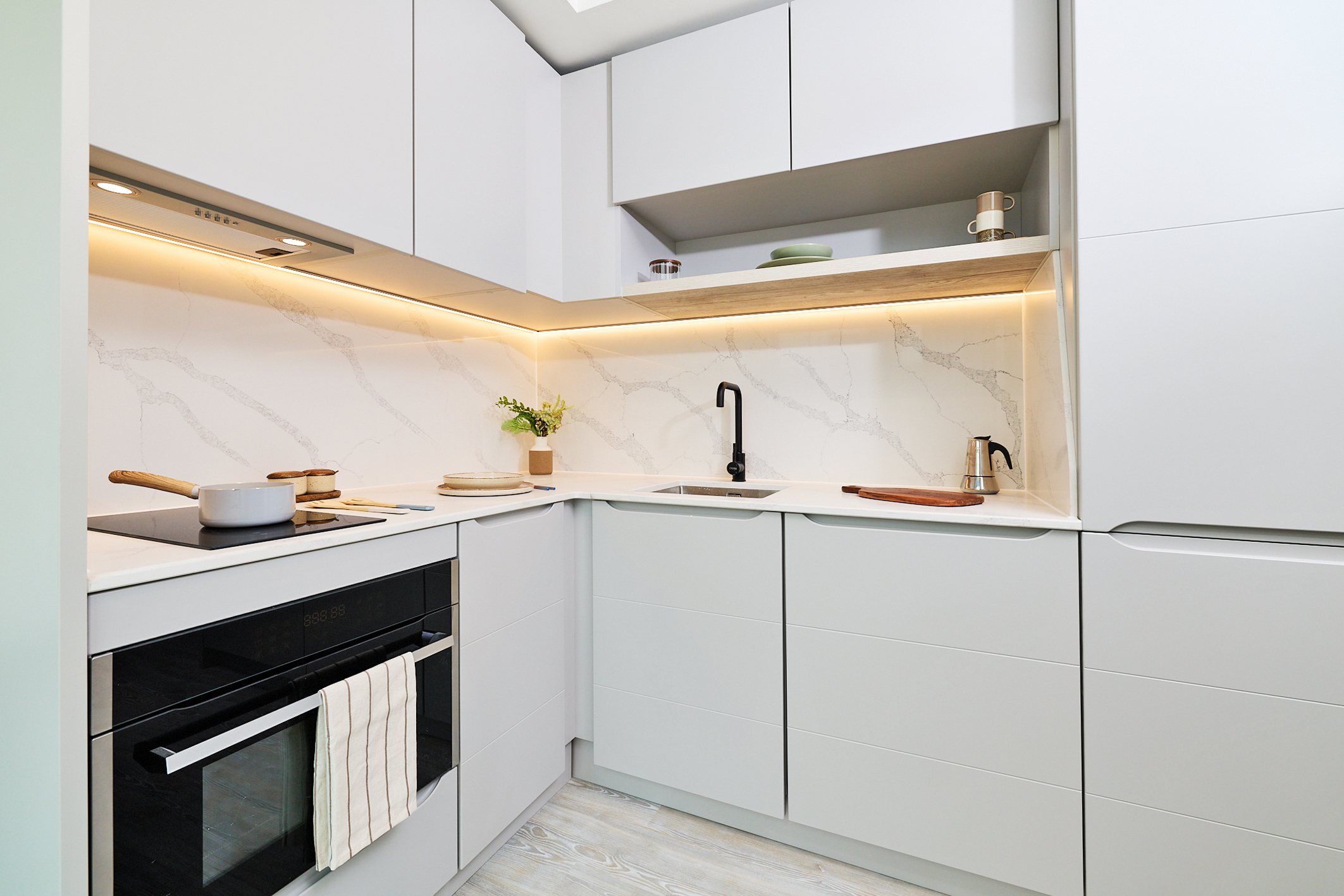 Modern white kitchen on Gough Street with marble backsplash, black faucet, induction stove, oven, and open shelves. Under-cabinet lighting highlights the minimalist design. A pot, pan, towel, and small plant are on the counter.