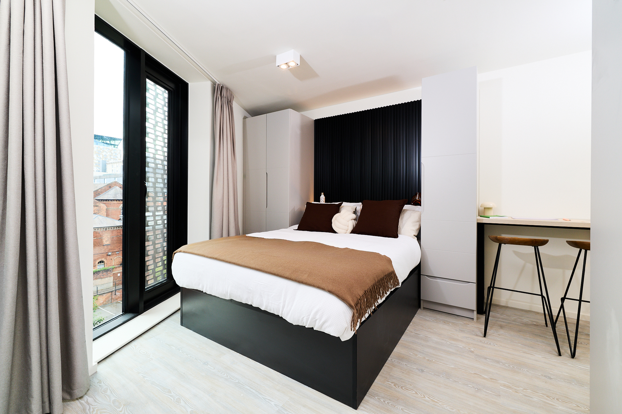 A modern Gough Street bedroom with a double bed, brown blanket, black headboard, two wardrobes, tall windows with curtains, bar stools, and a small counter. Light wooden flooring and city buildings visible outside.