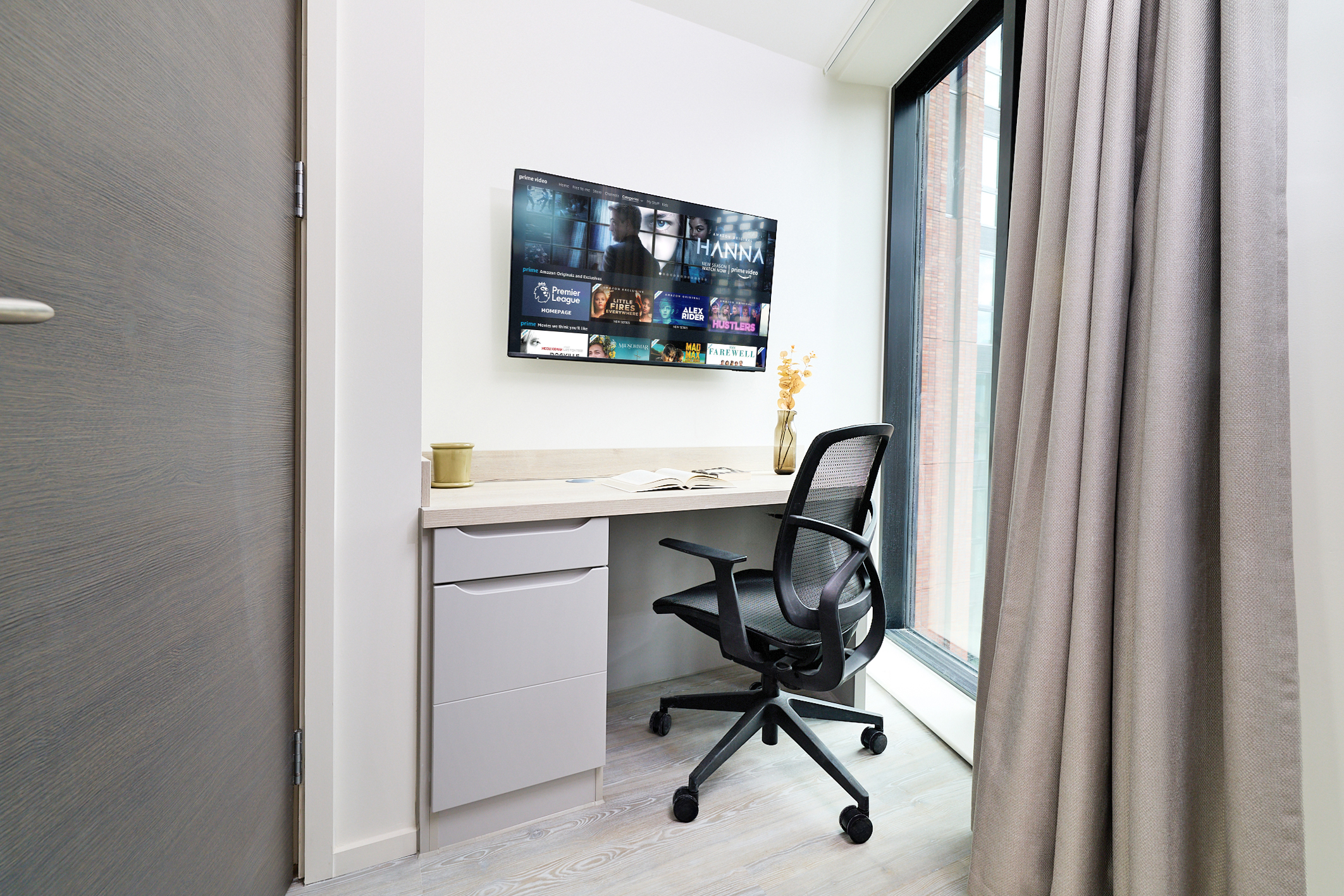 Minimalist home office on Gough Street featuring a desk, black office chair, wall-mounted TV displaying streaming apps, a cup, an open book, and a vase with dried flowers, situated next to a large window with gray curtains.