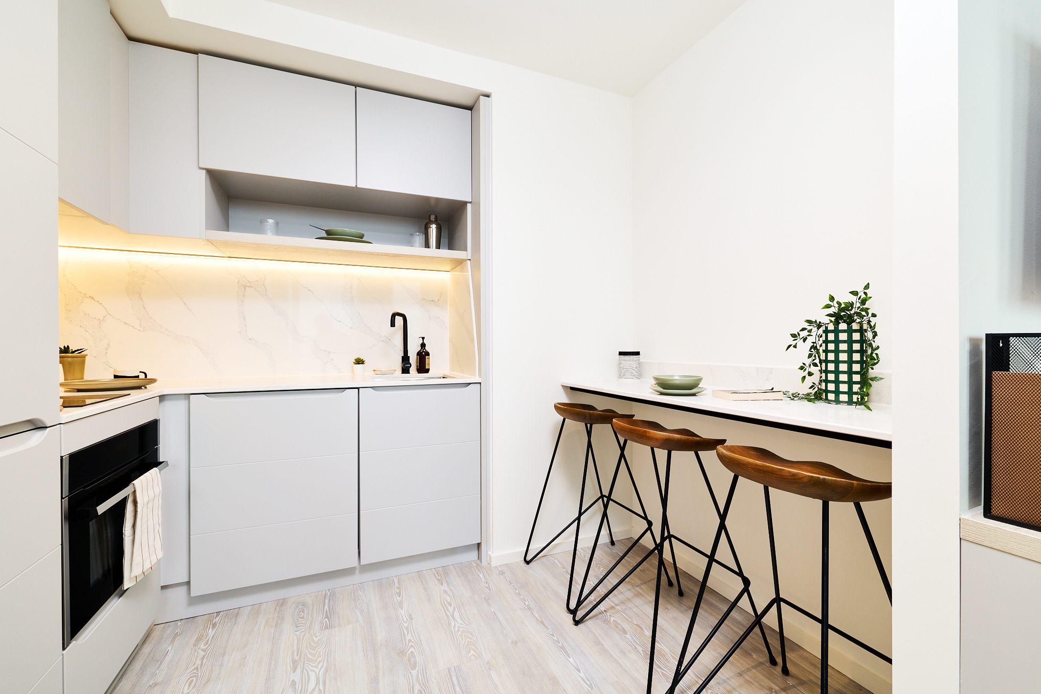 Modern minimalist kitchen on Gough Street with light gray cabinets, marble backsplash, black fixtures, and a breakfast bar with three wooden stools. Greenery decorates the counter. The space is clean and well-lit with under-cabinet lighting.