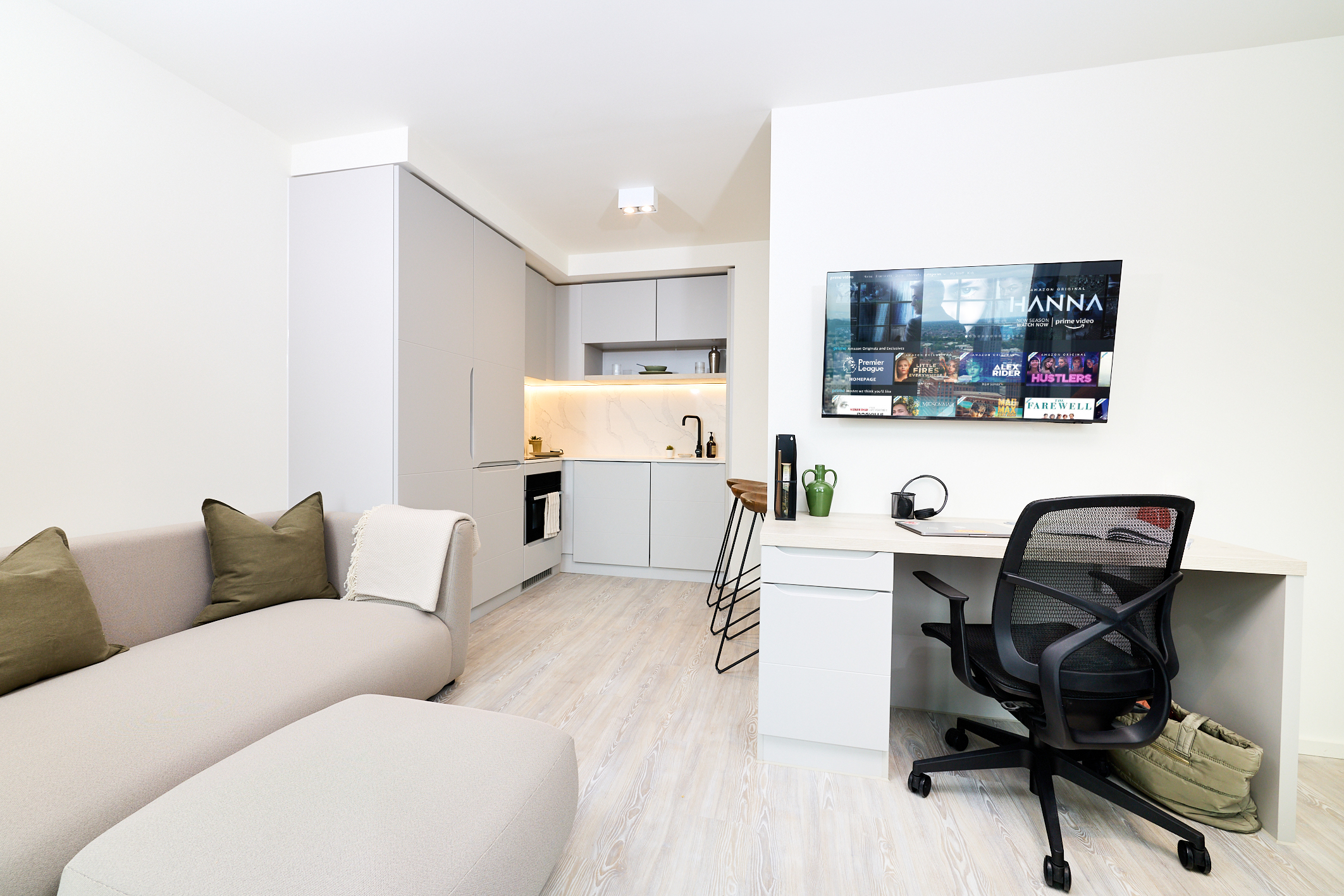 A modern studio apartment on Gough Street features a small kitchen, a beige sofa with green pillows, a desk with a chair, and a TV mounted on the wall displaying streaming options.
