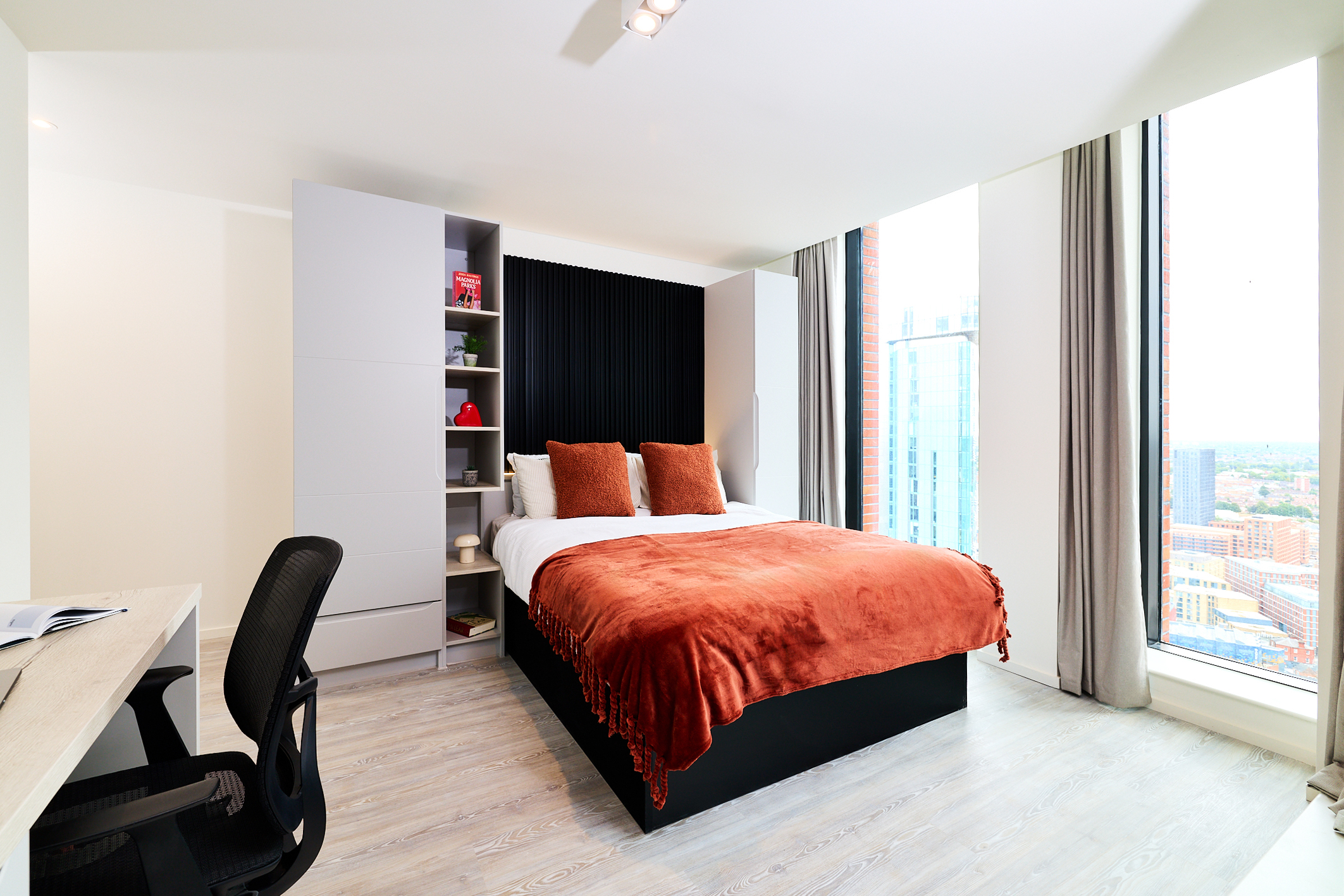 A modern bedroom on Gough Street features a double bed with orange bedding, two pillows, built-in shelves, a desk with a chair, and large floor-to-ceiling windows offering city views framed by light gray curtains.
