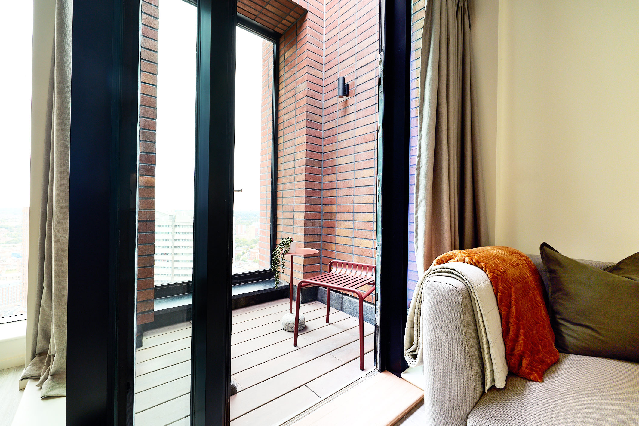 A small Gough Street balcony with red brick walls, a glass door, a red metal table and chair, and a potted plant. Inside, a beige sofa with green pillows and an orange blanket is visible.